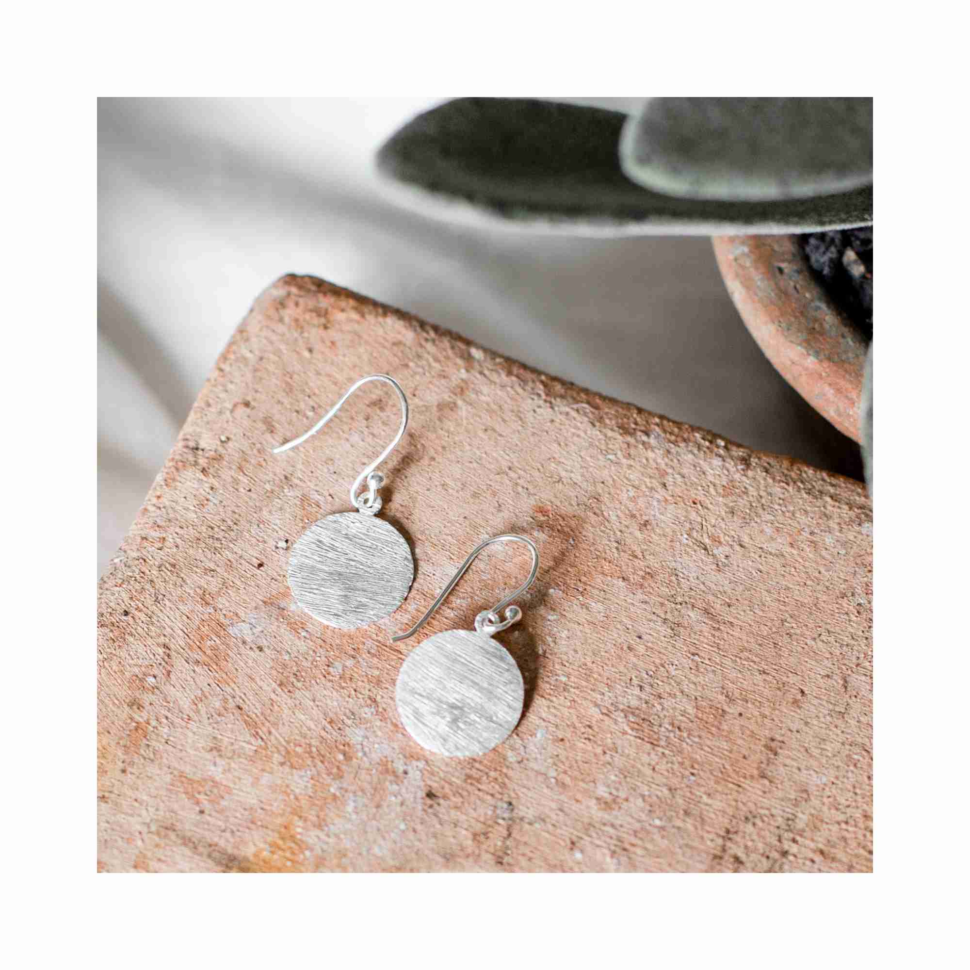 Lifestyle photoshoot of Shimmering Disks minimalist medium coin drop earrings in 92.5 recycled silver, featuring a hand-brushed matte finish, artistically displayed on a rustic terracotta brick with a succulent plant background.
