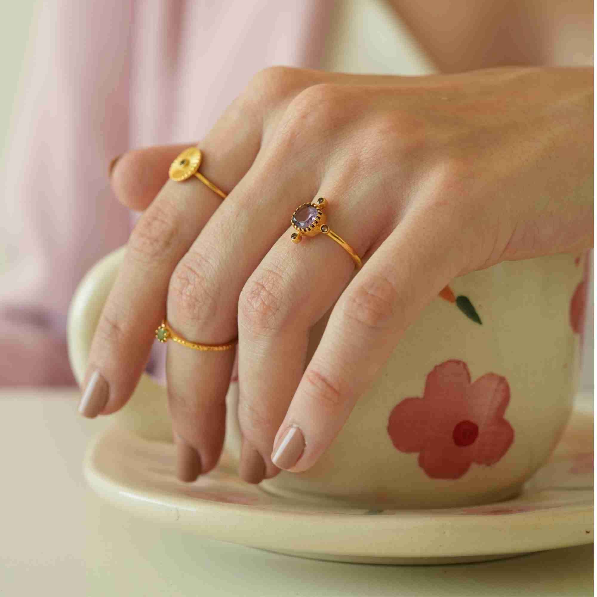A medium lifestyle shot of a model dressed in a pastel pink blazer holding a floral teacup and saucer, beautifully showcasing the Delicate Vintage Fluted Disc Ring in Gold by Protsaah Zurich as part of a chic, everyday editorial look.