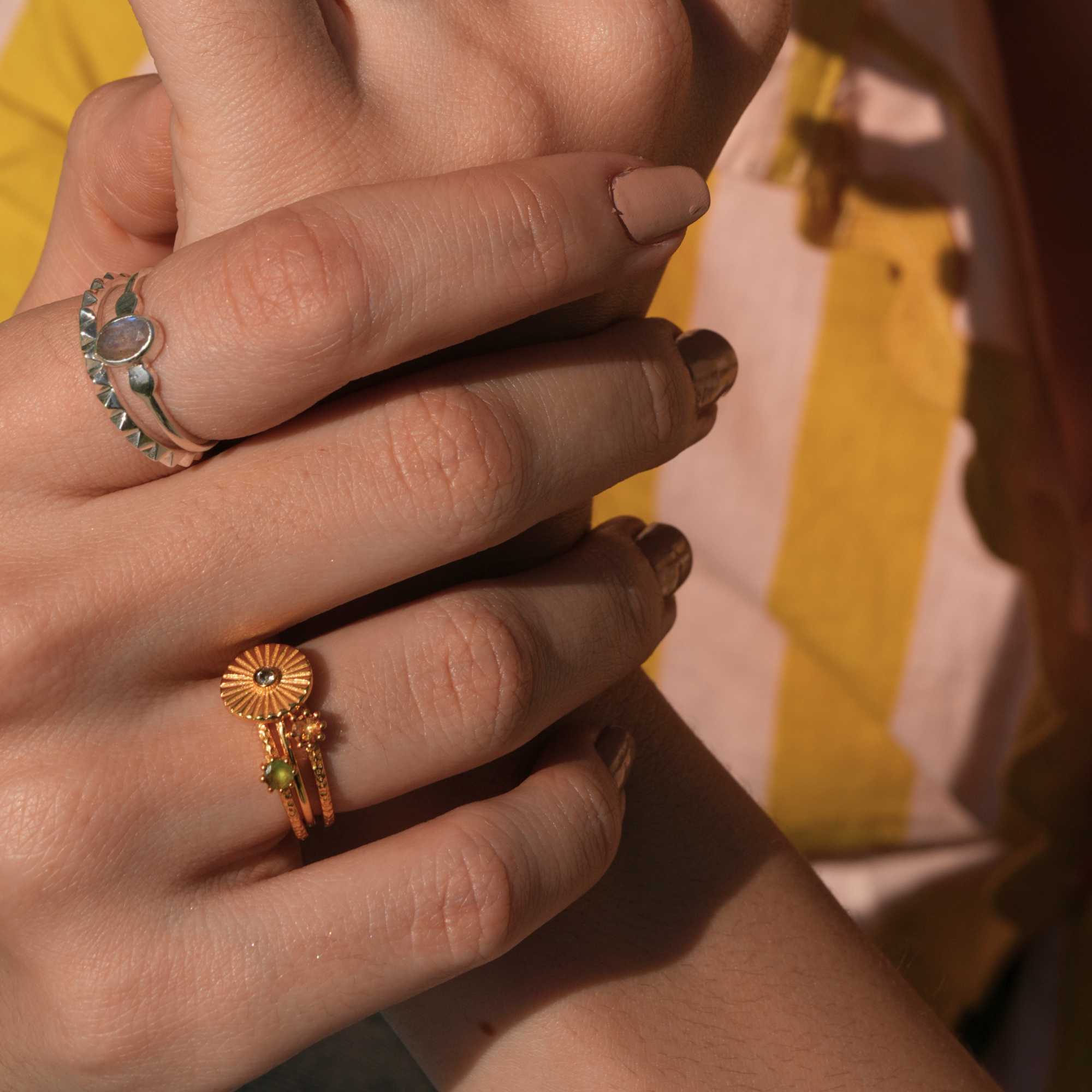 A close-up view of a model's hands with a beige manicure, showcasing a mixed metal ring stack featuring a silver gemstone ring alongside the Delicate Vintage Iolite Fluted Medallion Coin Ring in Gold by Protsaah Zurich.