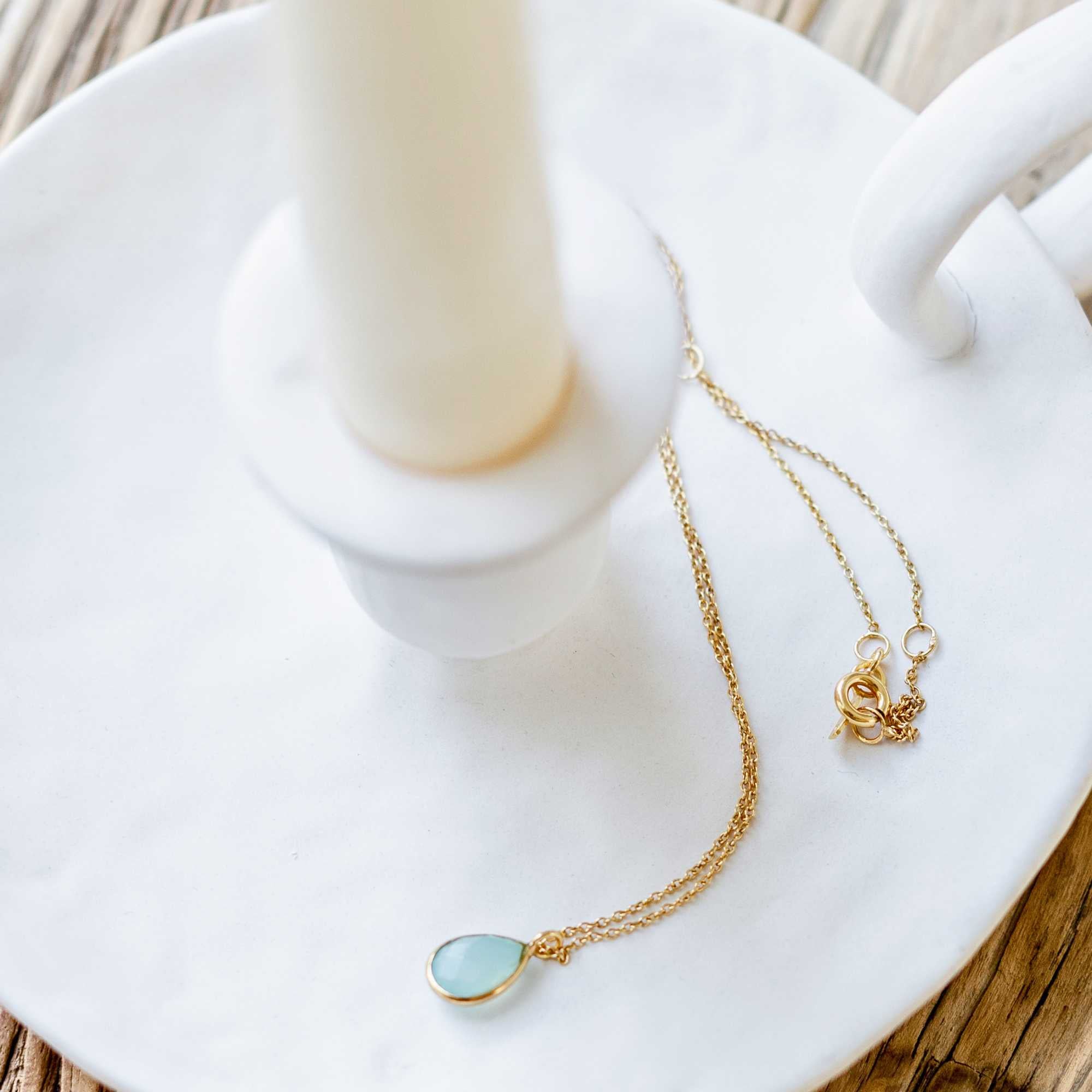 A gold pendant necklace featuring a delicate, bezel-set teardrop aqua chalcedony gemstone charm on a fine chain, shown both displayed on a white ceramic candlestick holder and worn by a model.