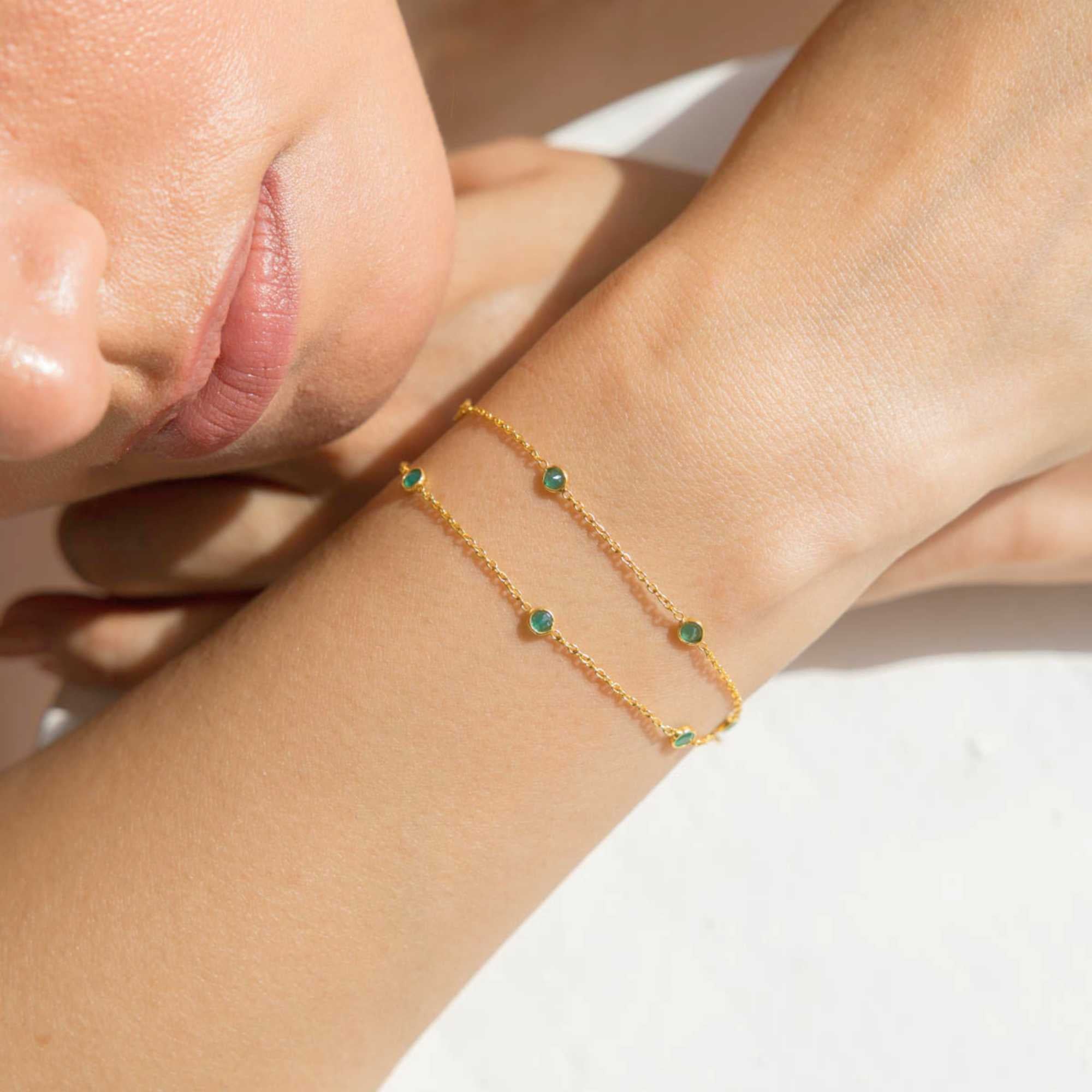A close-up lifestyle shot of a person's wrist wearing the Dew Drops Green Onyx Studded Chain Bracelet Gold, showcasing the delicate double gold chains and bezel-set green onyx gemstones under warm, natural sunlight.