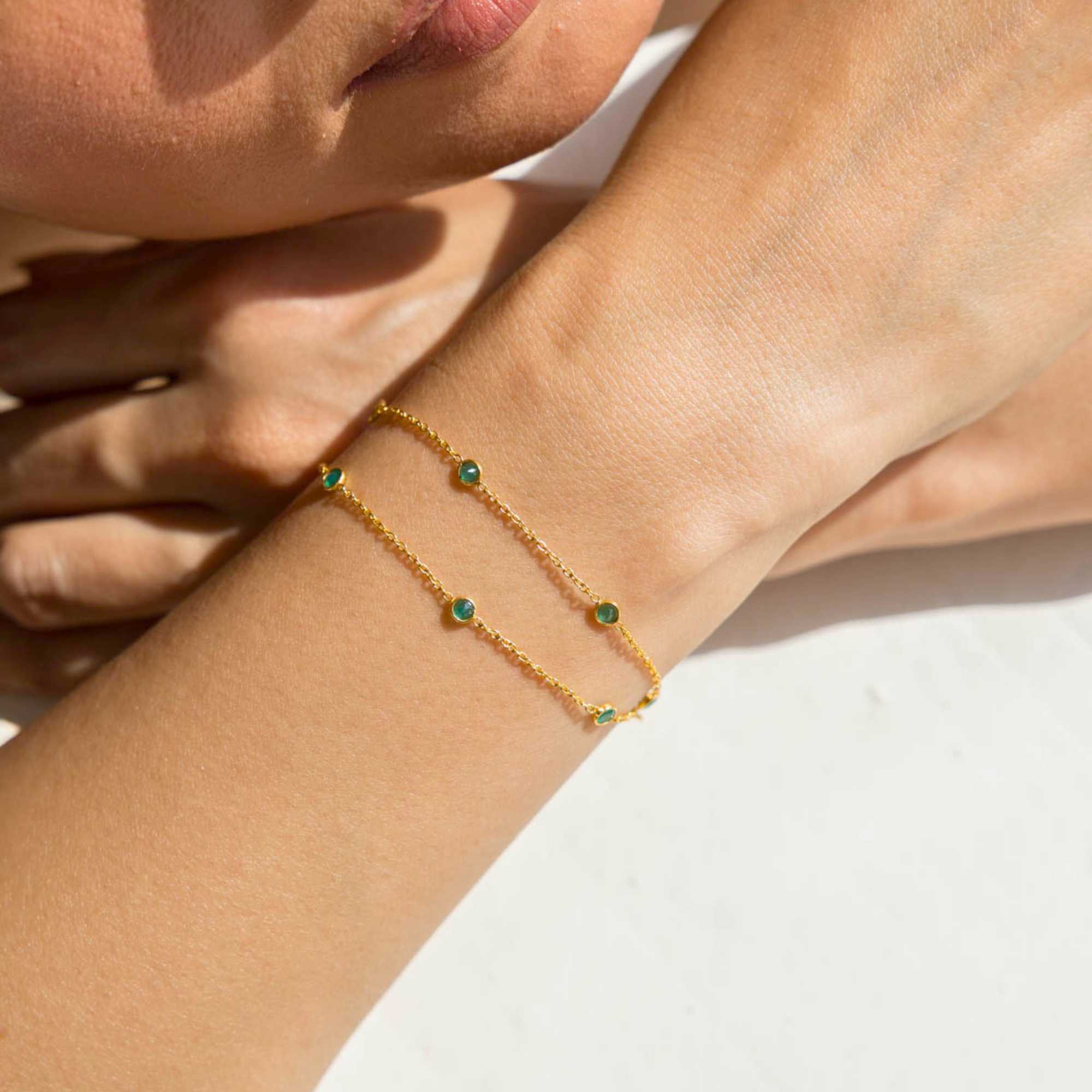 A close-up lifestyle shot of a person's wrist wearing the Dew Drops Green Onyx Studded Chain Bracelet Gold, showcasing the delicate double gold chains and bezel-set green onyx gemstones under warm, natural sunlight.