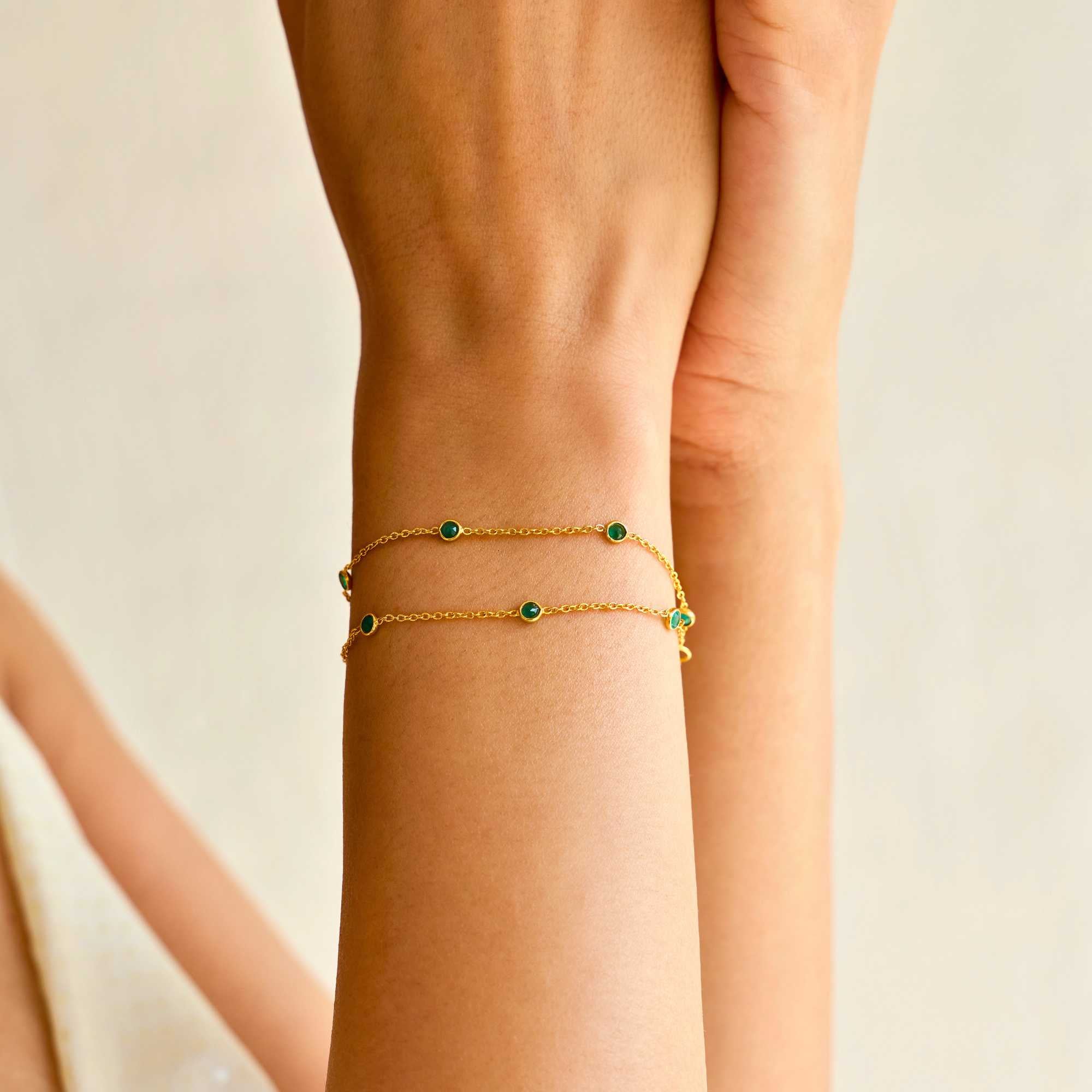 A close-up lifestyle shot of a person's wrist wearing the Dew Drops Green Onyx Studded Chain Bracelet Gold, showcasing the delicate double gold chains and bezel-set green onyx gemstones under warm, natural sunlight.