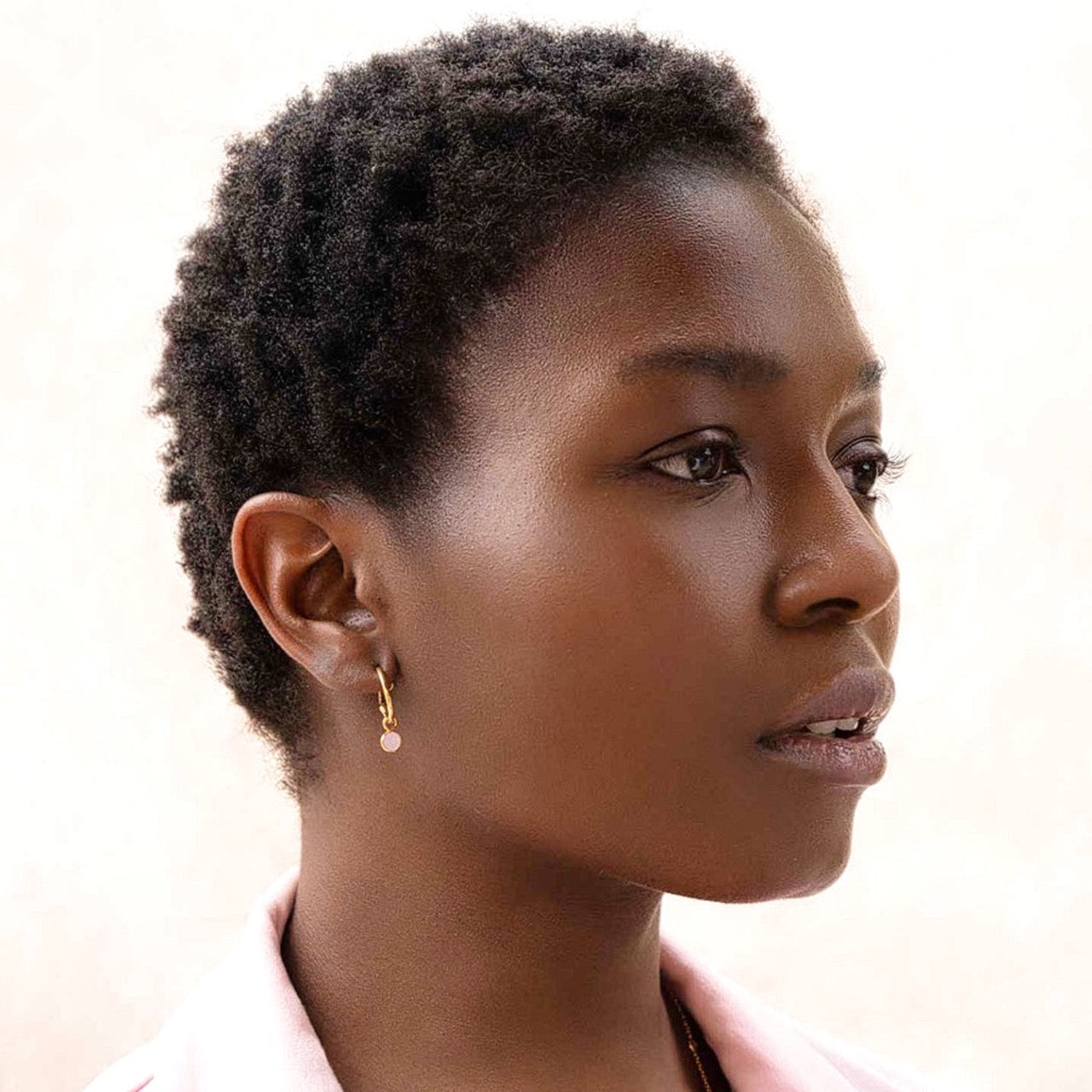 Close-up portrait campaign picture named Dew Drops showing a short-haired model in profile, showcasing the Hoop Earrings with Pink Chalcedony Charm Gold worn in her earlobe. The handcrafted 22K gold vermeil mini hoop features a small, dangling bezel-set round translucent pastel pink Chalcedony gemstone charm by Protsaah Zurich.