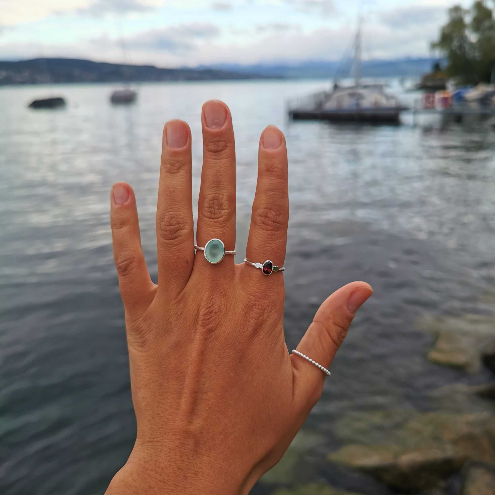 A detailed view of the Dew Drops Oval Aqua Chalcedony Adjustable Ring in sterling silver, showcased on a model's hand against a scenic, blurred lake background. The ring features a smooth, light-blue oval aqua chalcedony gemstone bezel-set on a textured silver band, layered gracefully alongside a dark garnet gemstone ring and a beaded silver band.