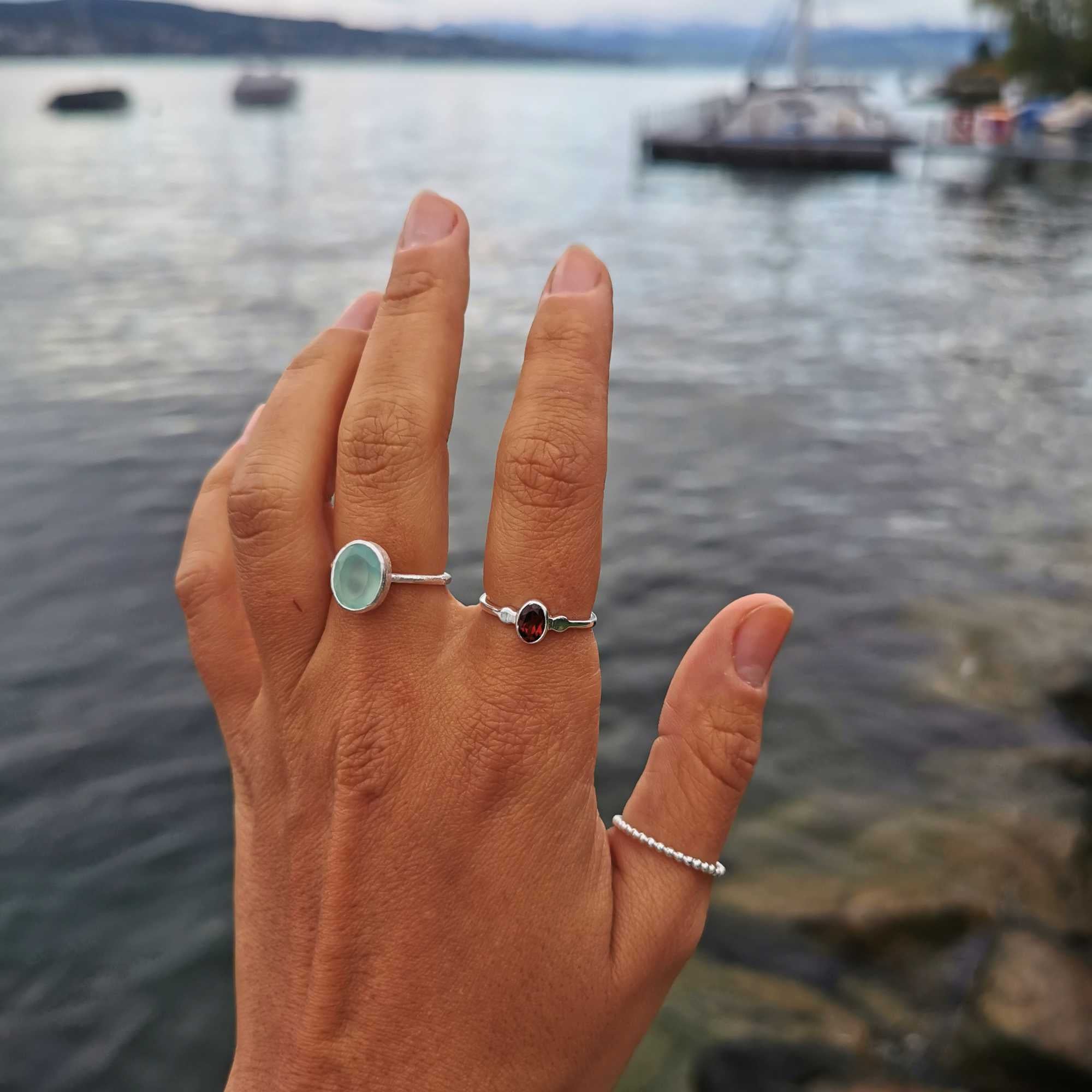 A detailed view of the Dew Drops Oval Aqua Chalcedony Adjustable Ring in sterling silver, showcased on a model's hand against a scenic, blurred lake background. The ring features a smooth, light-blue oval aqua chalcedony gemstone bezel-set on a textured silver band, layered gracefully alongside a dark garnet gemstone ring and a beaded silver band.