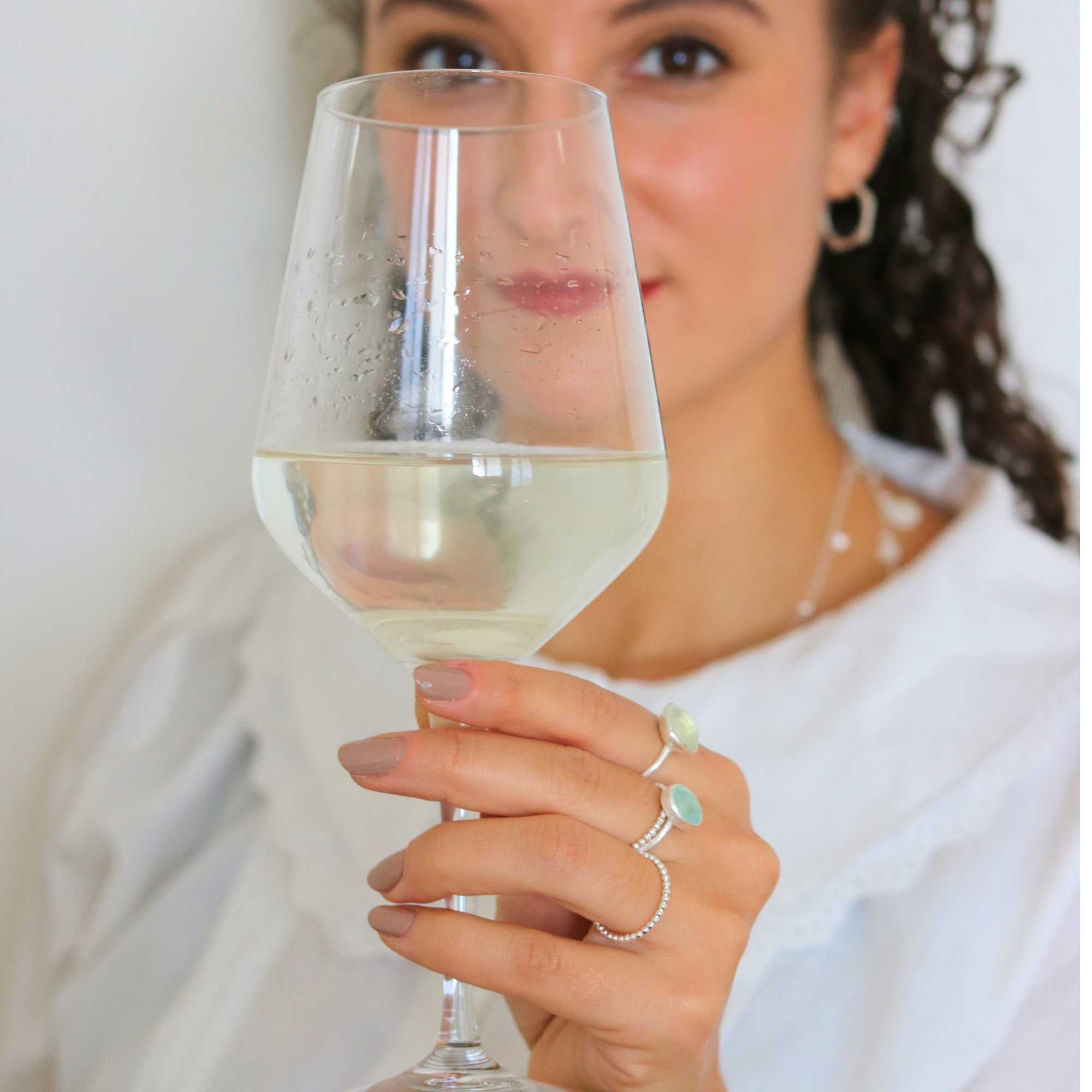 A detailed view of the Dew Drops Oval Aqua Chalcedony Adjustable Ring in sterling silver, showcased on a model's hand against a scenic, blurred lake background. The ring features a smooth, light-blue oval aqua chalcedony gemstone bezel-set on a textured silver band, layered gracefully alongside a dark garnet gemstone ring and a beaded silver band.