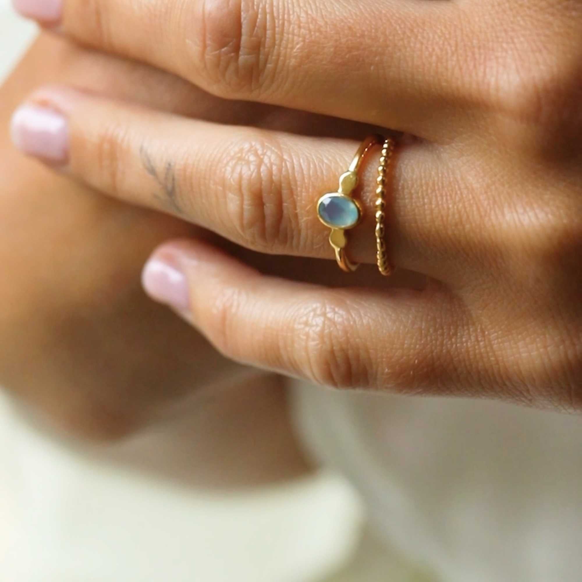 A gold adjustable ring featuring a small, bezel-set oval aqua chalcedony gemstone at its center, flanked by delicate metallic droplet accents on a smooth gold band.