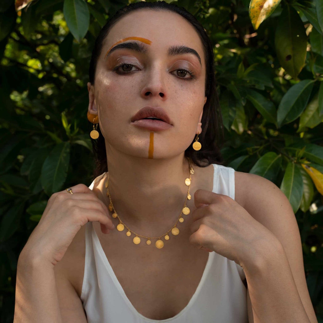 Artistic editorial photoshoot of Shimmering Disks large gold minimalist coin drop earrings and matching gold disk necklace, styled on a model with sunset-orange avant-garde makeup against a natural green foliage background to showcase the 22K gold vermeil luster.