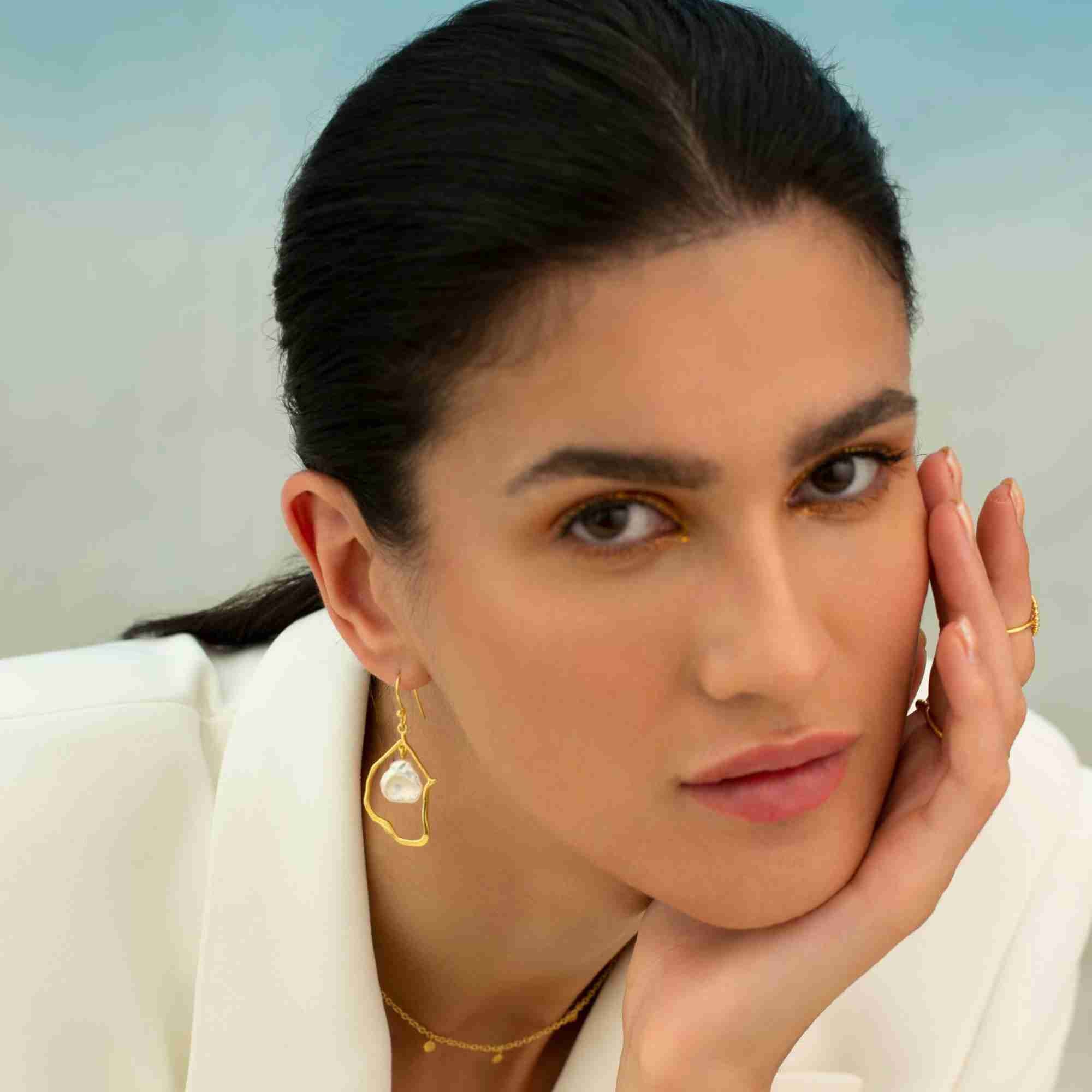 Editorial portrait of a dark-haired model wearing a white blazer, posing with her hand to her face to showcase the gold baroque pearl drop statement earring.