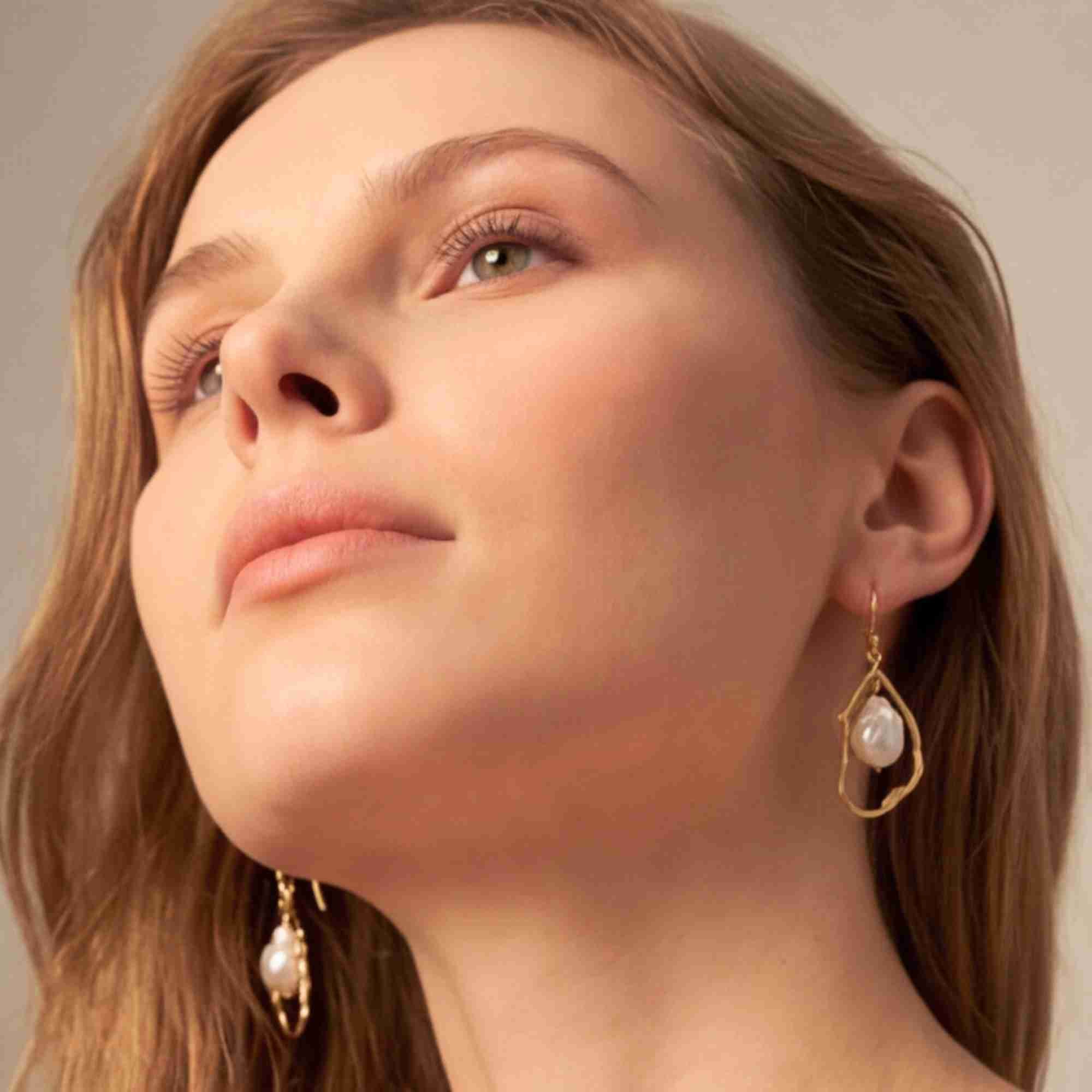 Low-angle studio portrait of a model looking upward, displaying the gold baroque pearl drop statement earrings on both ears.