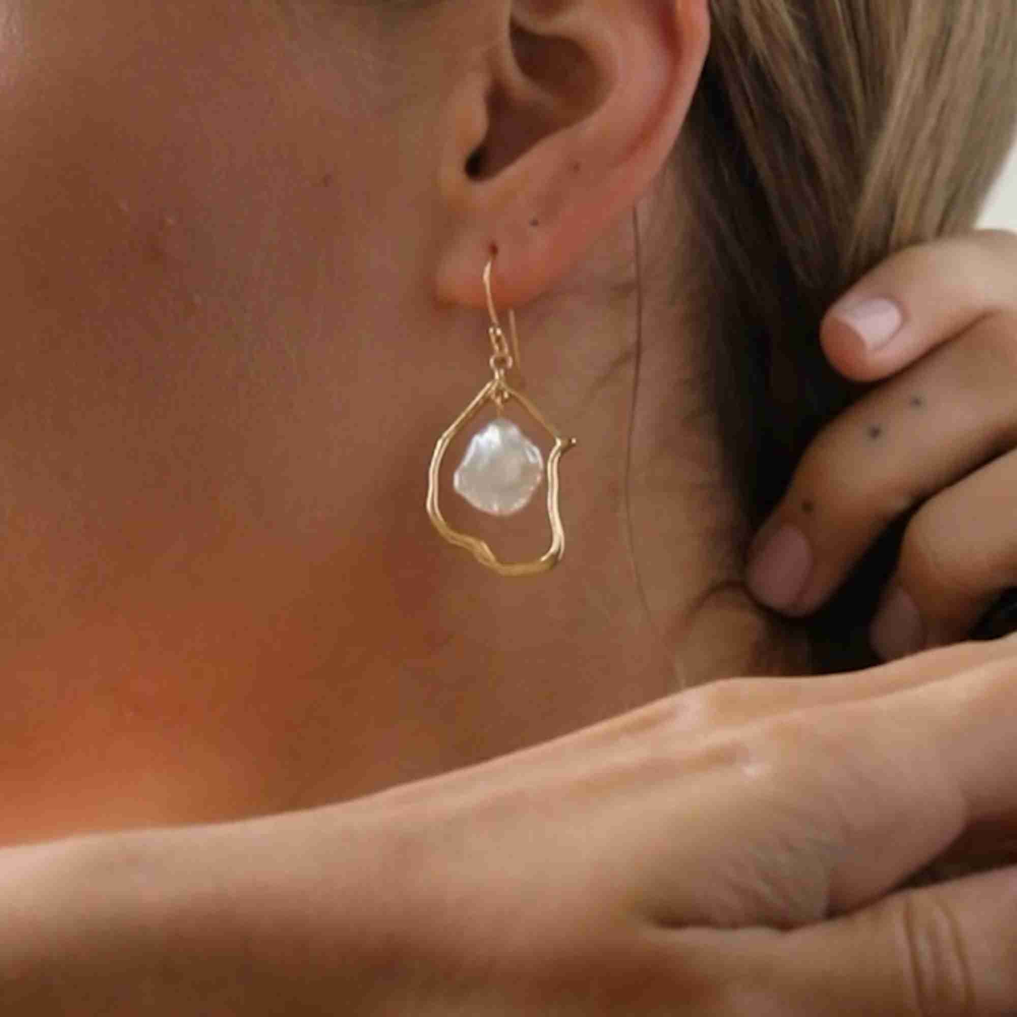 Close-up macro detail of the gold wire frame drop earring with a central floating baroque pearl dangling from an ear hook wire.