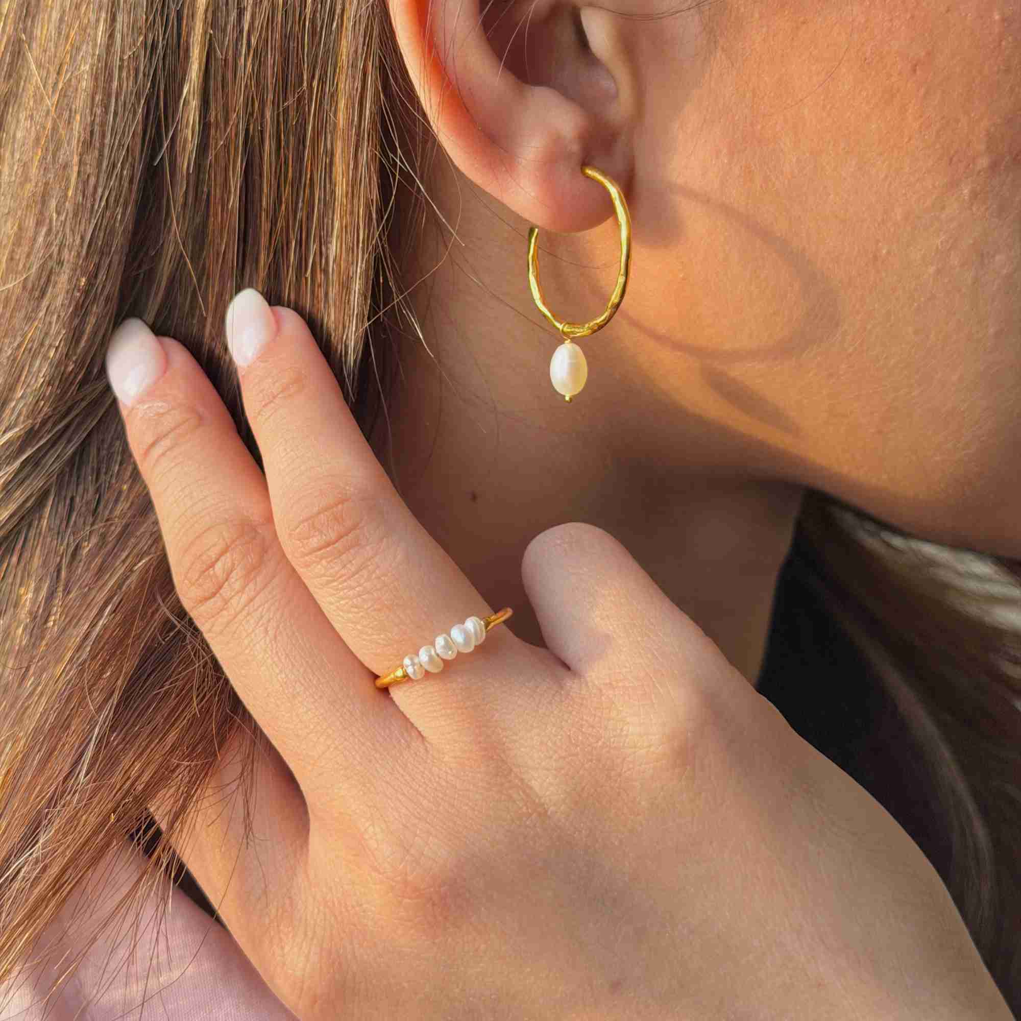 Warmly lit profile photo of a model placing her hand near her hair, displaying the gold PEARLS GALORE Linear Seed Pearl Bar Ring alongside a matching gold hoop pearl drop earring.