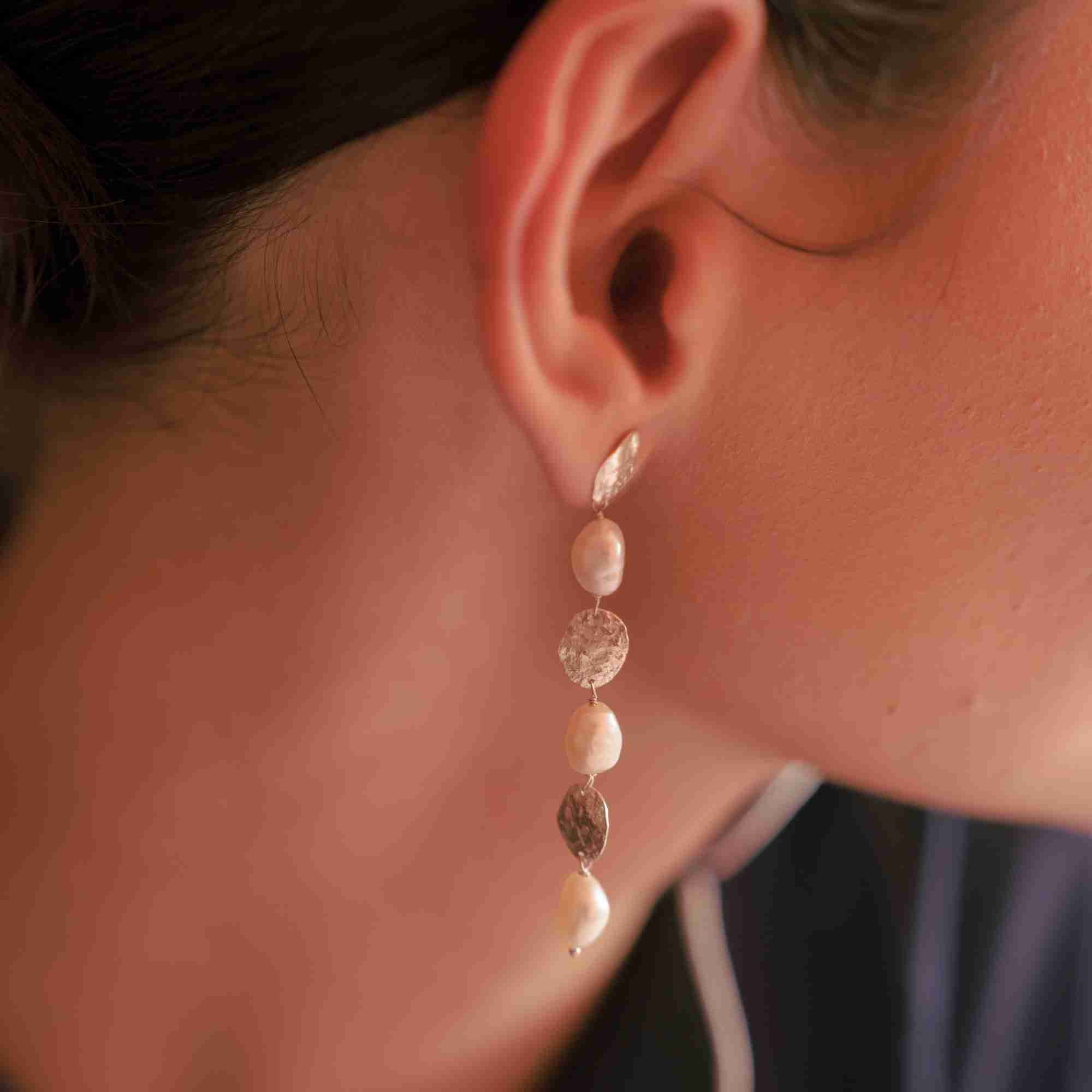 Warmly lit macro close-up of a model wearing the PEARLS GALORE Molten Gold & Pearl Cascade Statement Earrings, showing the alternating pattern of textured gold discs and organic pearls dangling in a long strand.