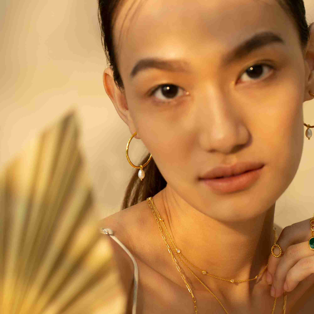 Warmly lit close-up portrait of a model wearing the gold PEARLS GALORE Teardrop Hoop Earrings with a small pearl charm dangling from each hoop.
