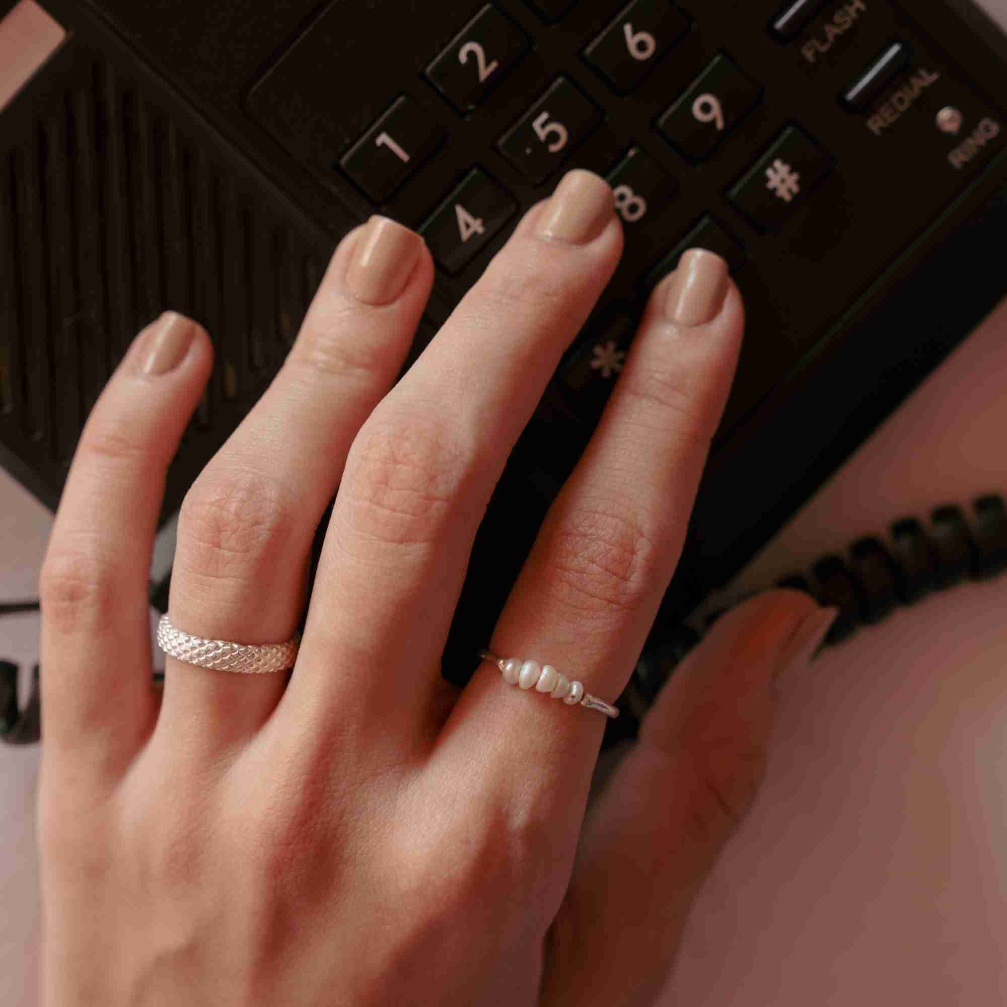 Top-down close-up of a model's hand with nude manicured nails pressing a vintage black landline telephone, featuring the silver Pearls Galore Linear Seed Pearl Bar Ring paired with a textured silver band.