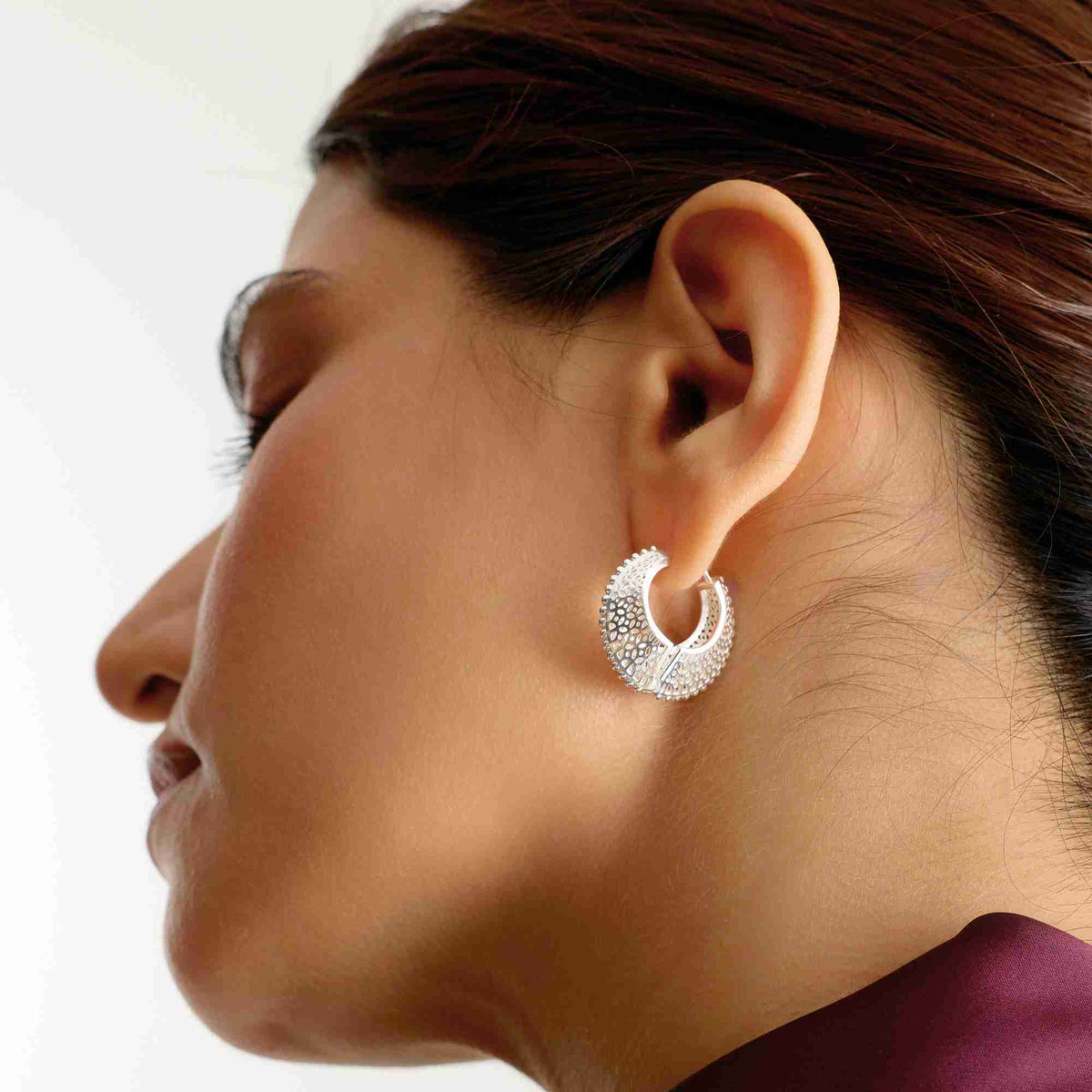 Side profile portrait of a model wearing a deep purple top, highlighting the intricate openwork filigree lace pattern on the sterling silver sculptural hoop earrings.