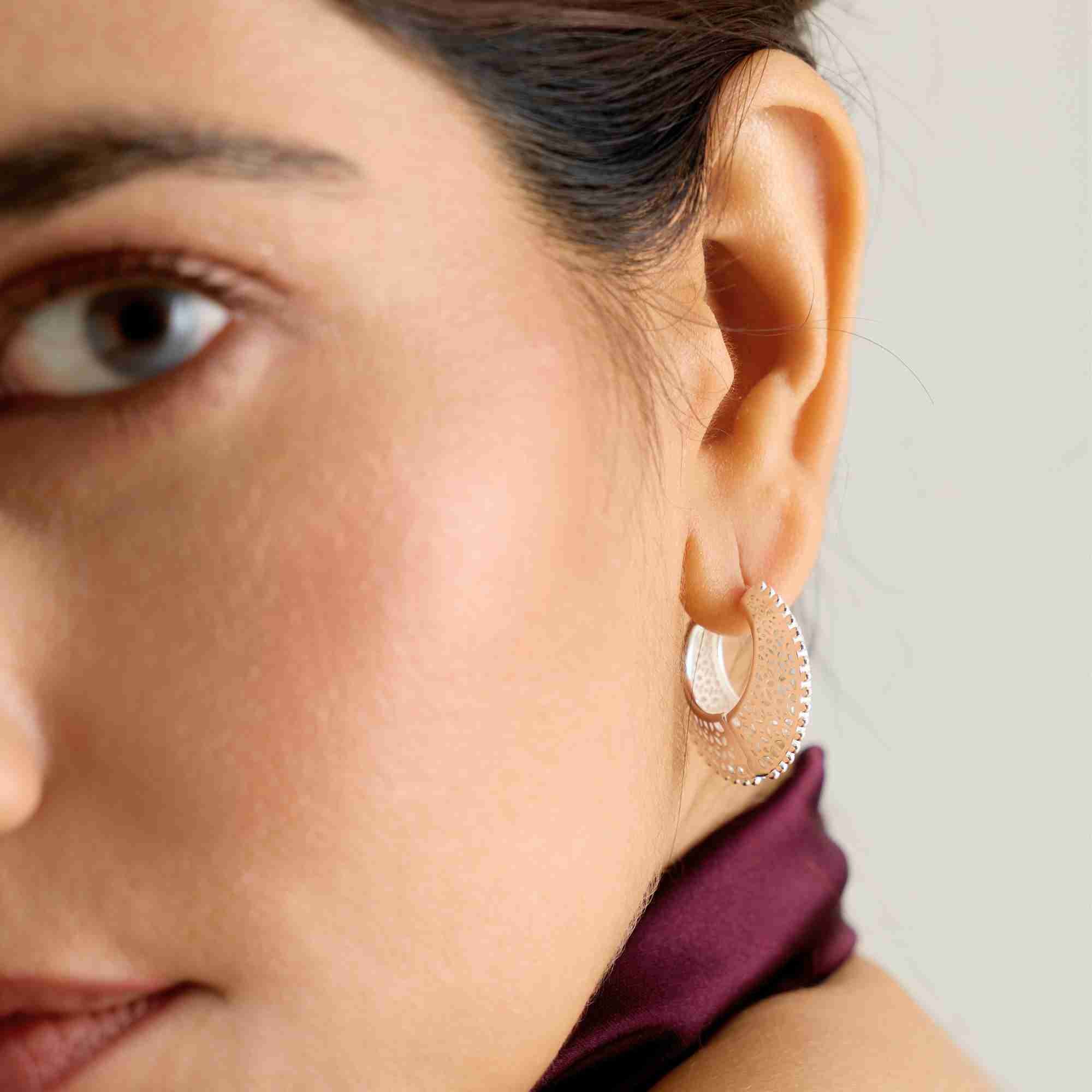 Close-up three-quarter view of a model wearing the silver Ribbons & Lace Filigree Sculptural Hoop Earrings paired with a dark silk top.