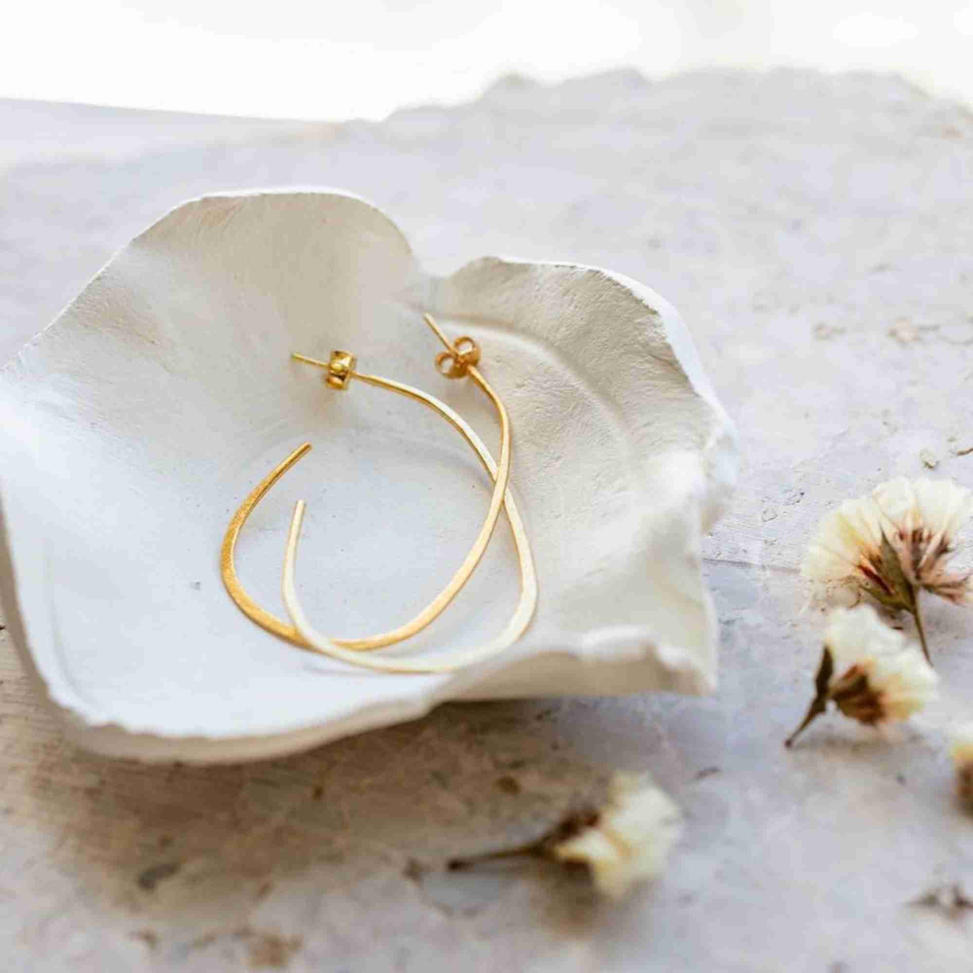 Flat lay photo of the gold Ribbons & Lace Organic Oval Fine Hoop Earrings resting inside a handmade white ceramic ring dish next to dried white blossoms.