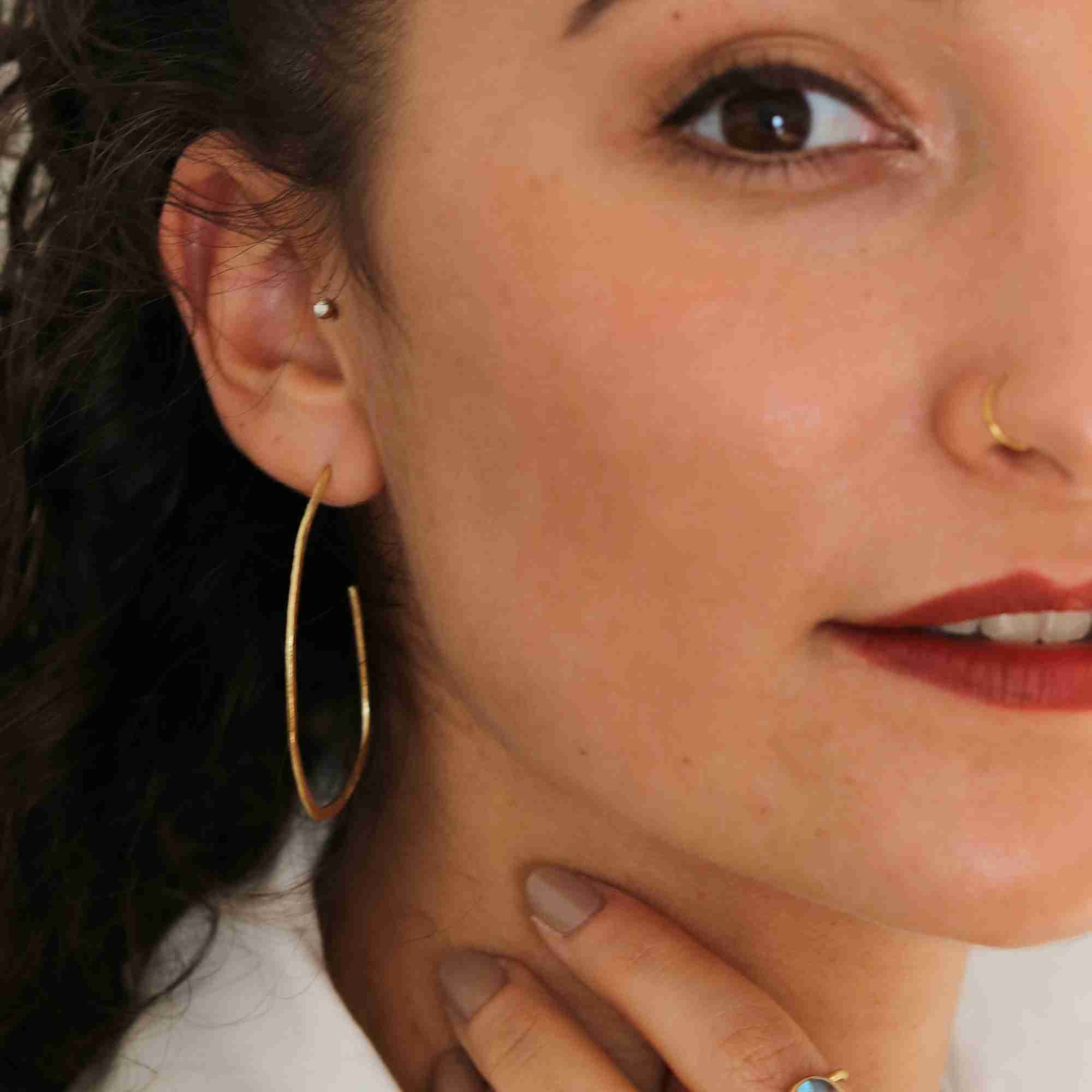 Close-up model view named Ribbon & Lace (3).jpg showing a woman wearing the Ribbons & Lace Organic Oval Fine Hoop Earrings Gold, highlighting the slender, elongated open oval shape and hand-brushed golden finish of the hoop resting alongside her jawline by Protsaah Zurich.
