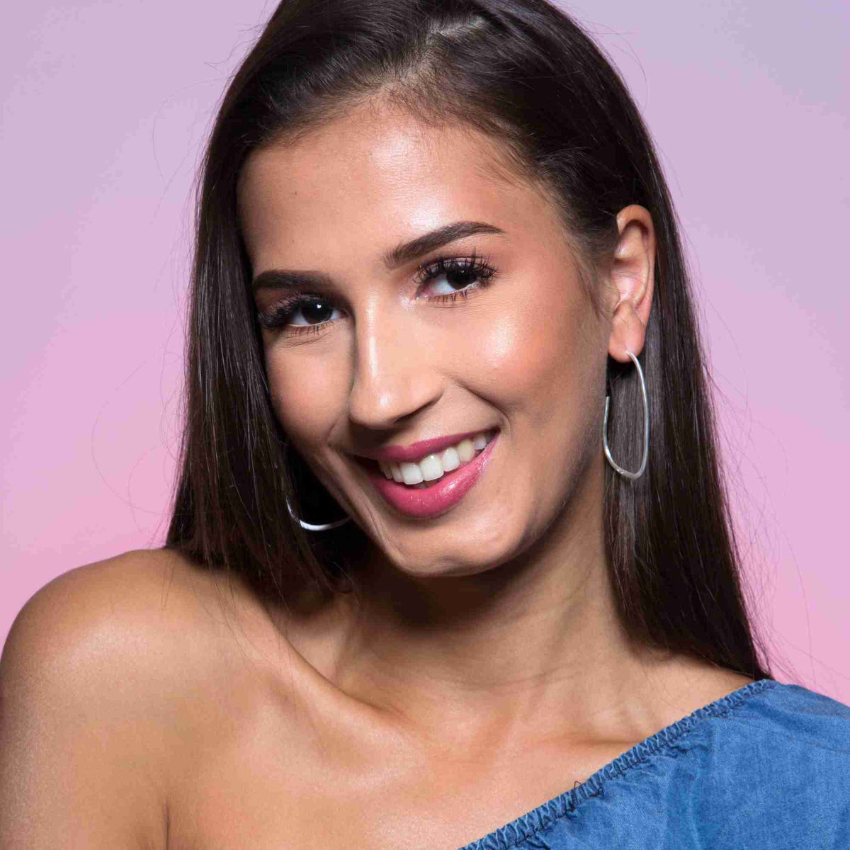 Smiling model against soft pink wearing fine organic oval hoop earrings in silver