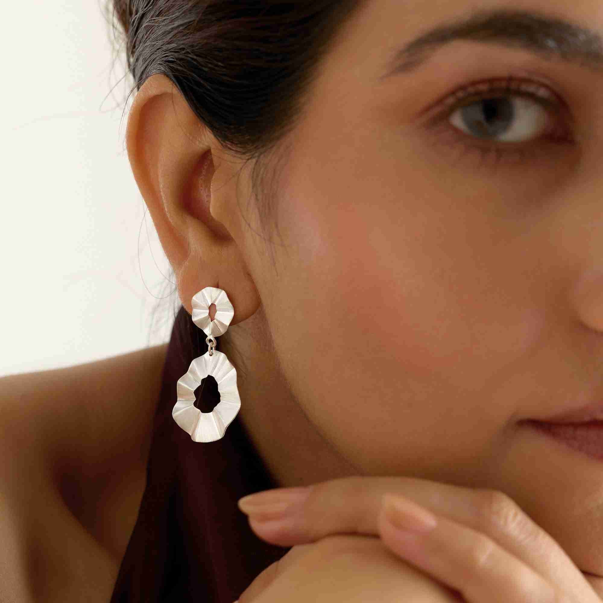 Lifestyle portrait view named Ribbon & Lace (12).jpg showing a model resting her chin on her hand while wearing the Ribbons & Lace Ruffle Fold Drop Statement Earrings Silver, highlighting the elegant drop and fine artisan texture of the sterling silver jewelry by Protsaah Zurich.