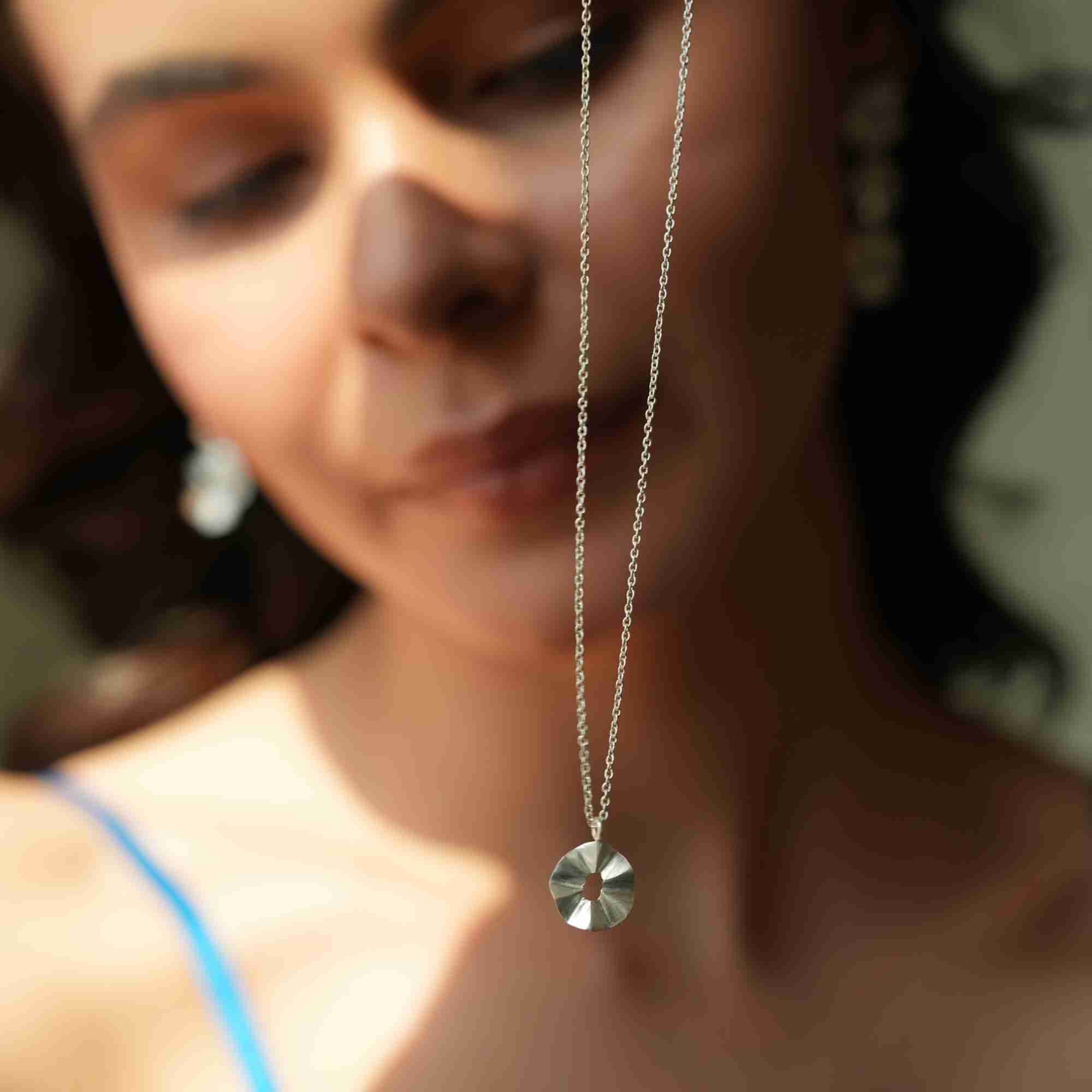 Close-up shot of the sterling silver Ribbons & Lace Ruffle Furls Pendant Necklace suspended in the foreground with a model softly blurred in the background.