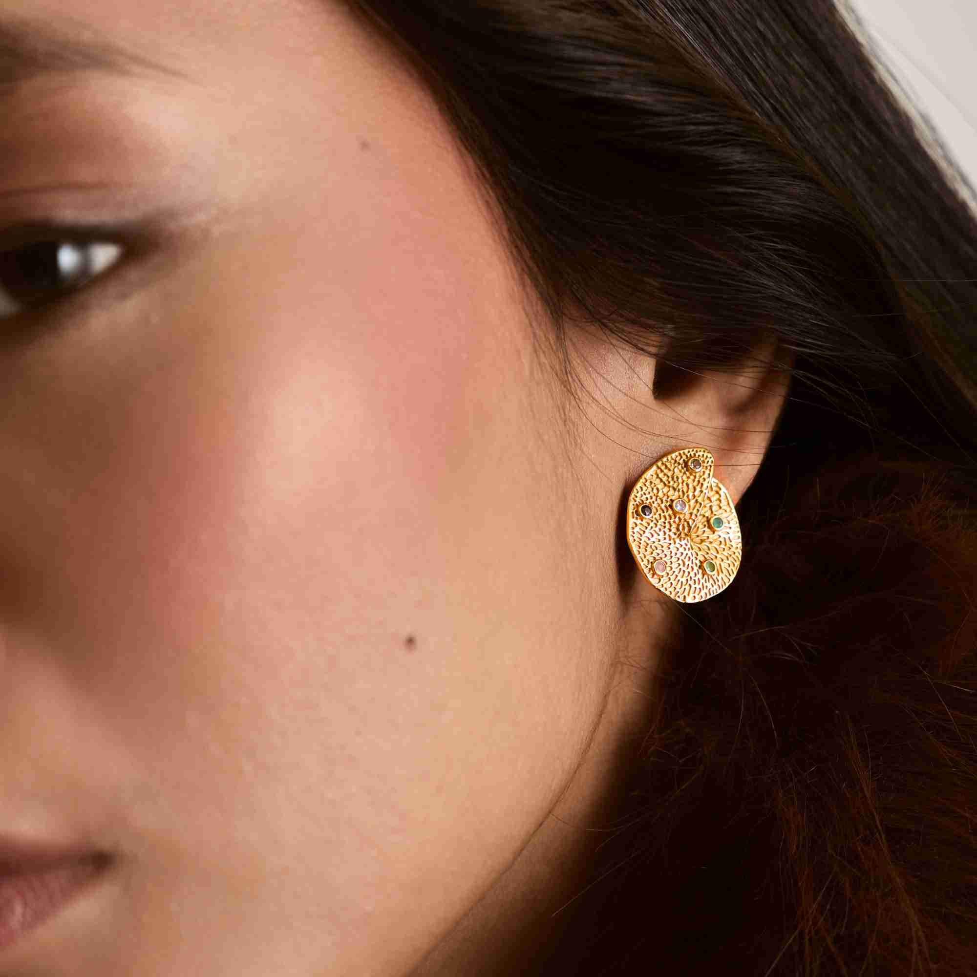 Close-up model view named Ribbon & Lace.jpg showing a woman wearing the Ribbons and Lace Galaxy Multi-Gemstone Stud Earrings Gold, showcasing the large, organically shaped gold disc with an intricate textured lace pattern studded with tiny, multi-colored embedded gemstones by Protsaah Zurich.