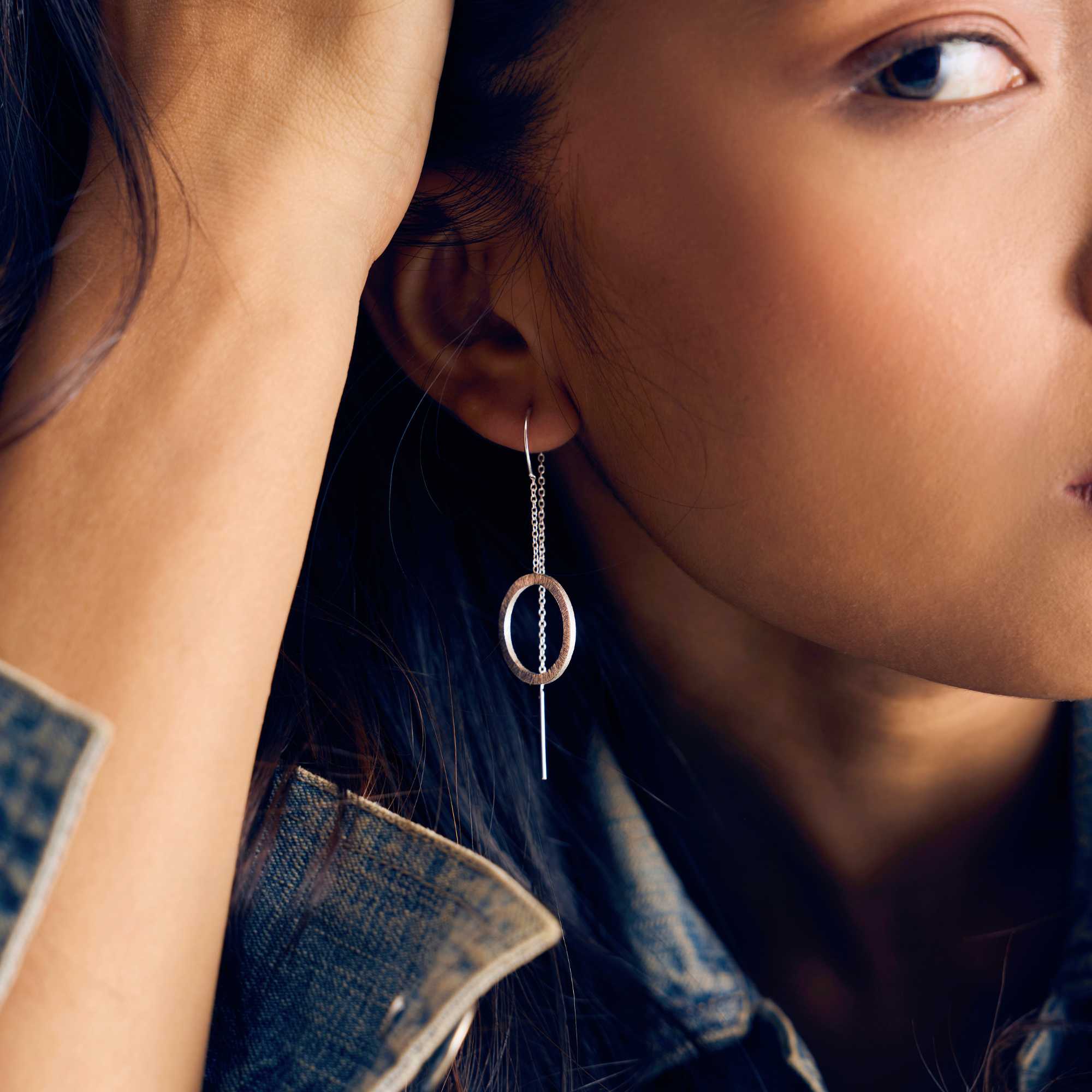 A model showcasing the Shimmering Disks Circle Chain Threader Earrings in silver, featuring a delicate fine drop chain that threads through the earlobe to suspend a textured, brushed metallic circular geometric charm.