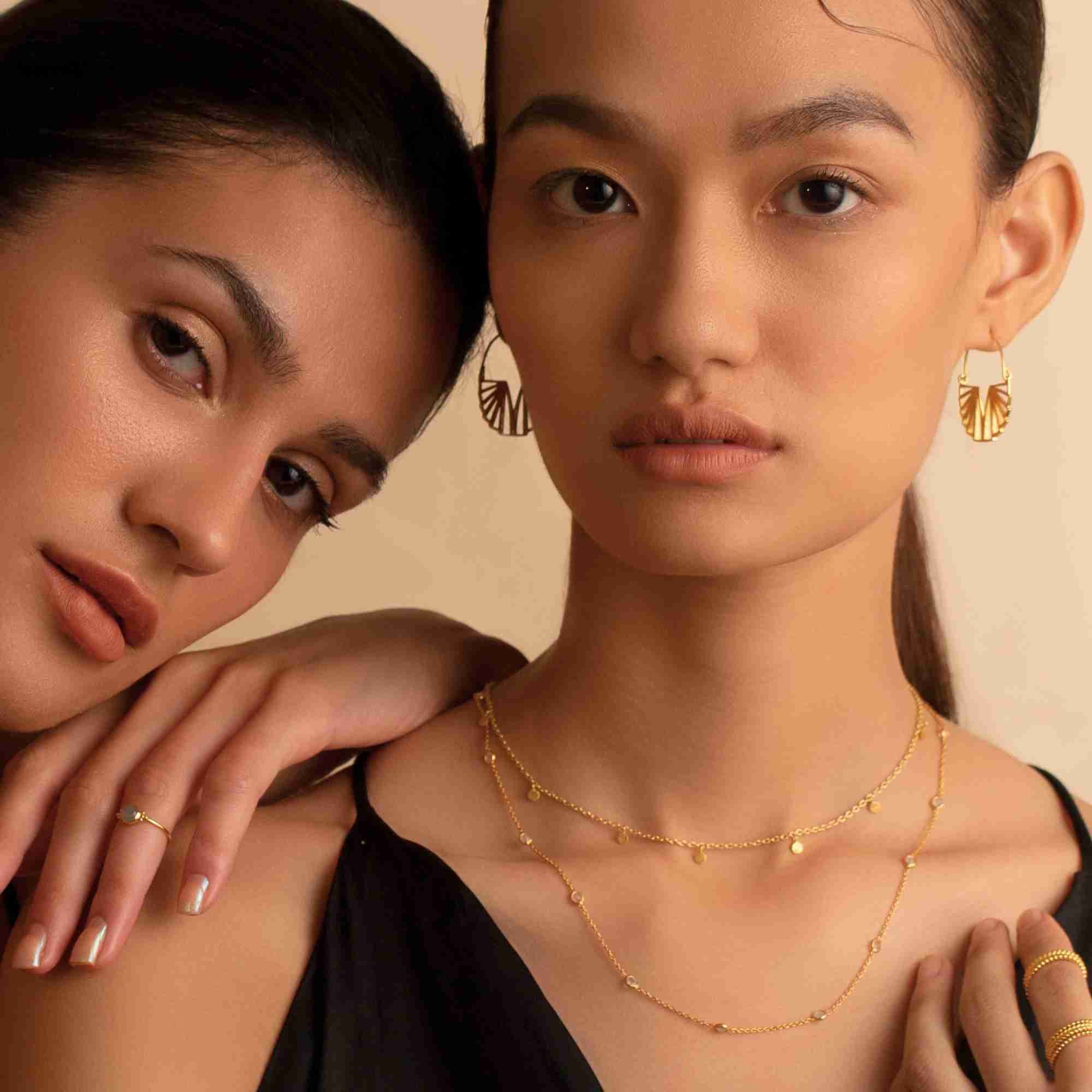 Warm studio campaign portrait of two models; the model on the right wears the gold Shimmering Disks Constellation Disc Chain Necklace layered with a longer fine bezel chain, styled with gold fan hoop earrings and gold rings.