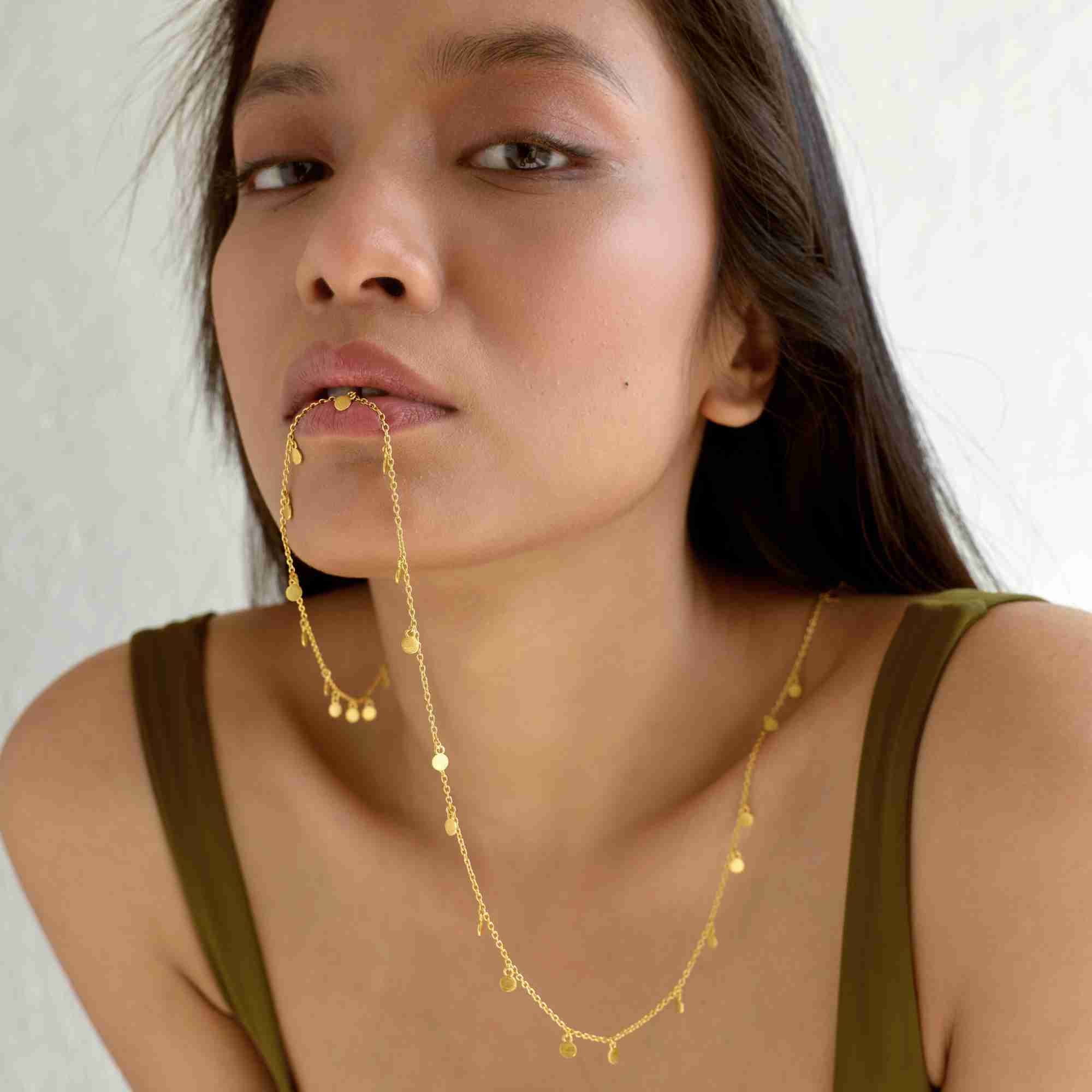 Close-up editorial shot of a model with the gold Shimmering Disks Constellation Disc Chain Necklace held gently between her lips.