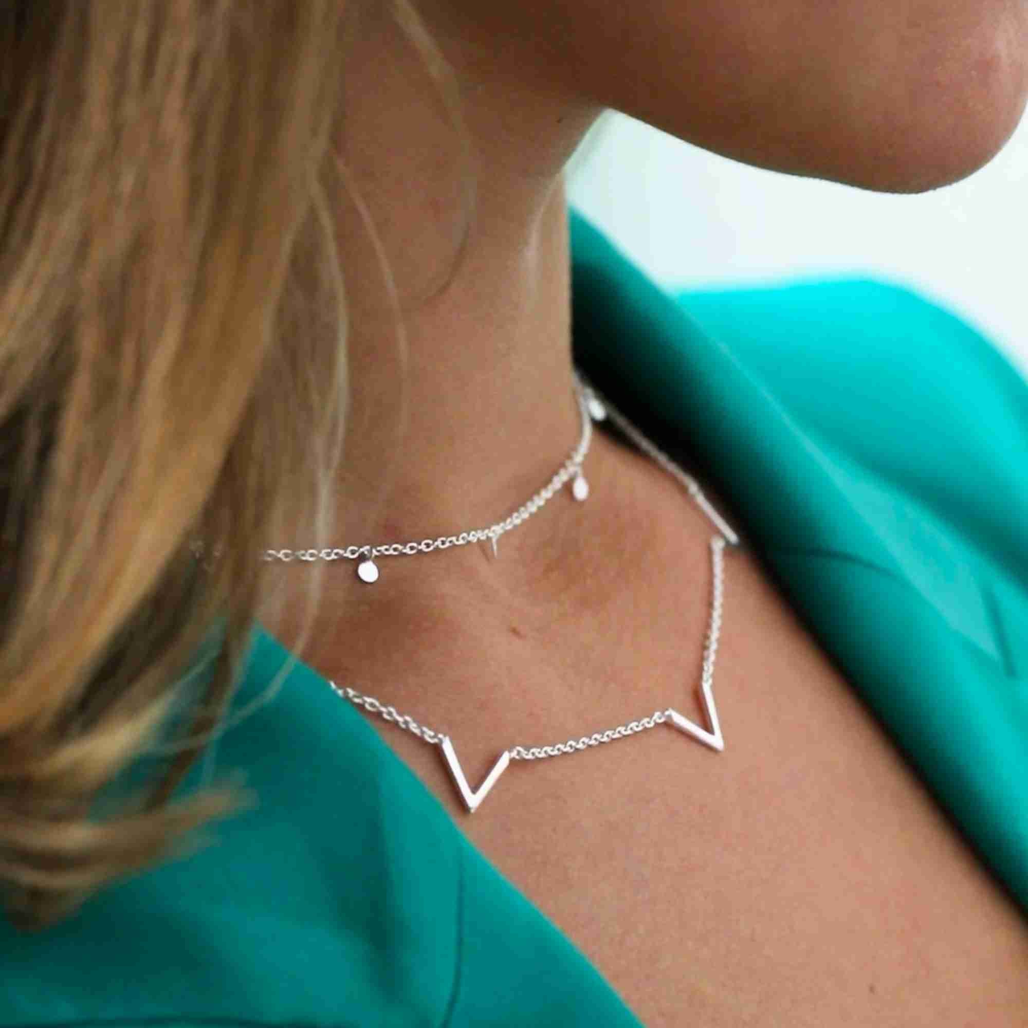 Model wearing the silver Shimmering Disks Constellation Disc Chain Necklace layered as a choker above a geometric chevron bar necklace, styled with a vibrant teal blazer.