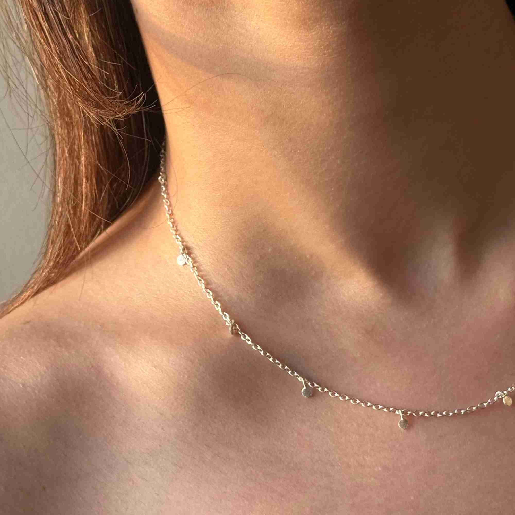 Angled close-up shot of a model wearing the silver Shimmering Disks Constellation Disc Chain Necklace, showing the tiny hanging coin charms resting against the collarbone under warm lighting.