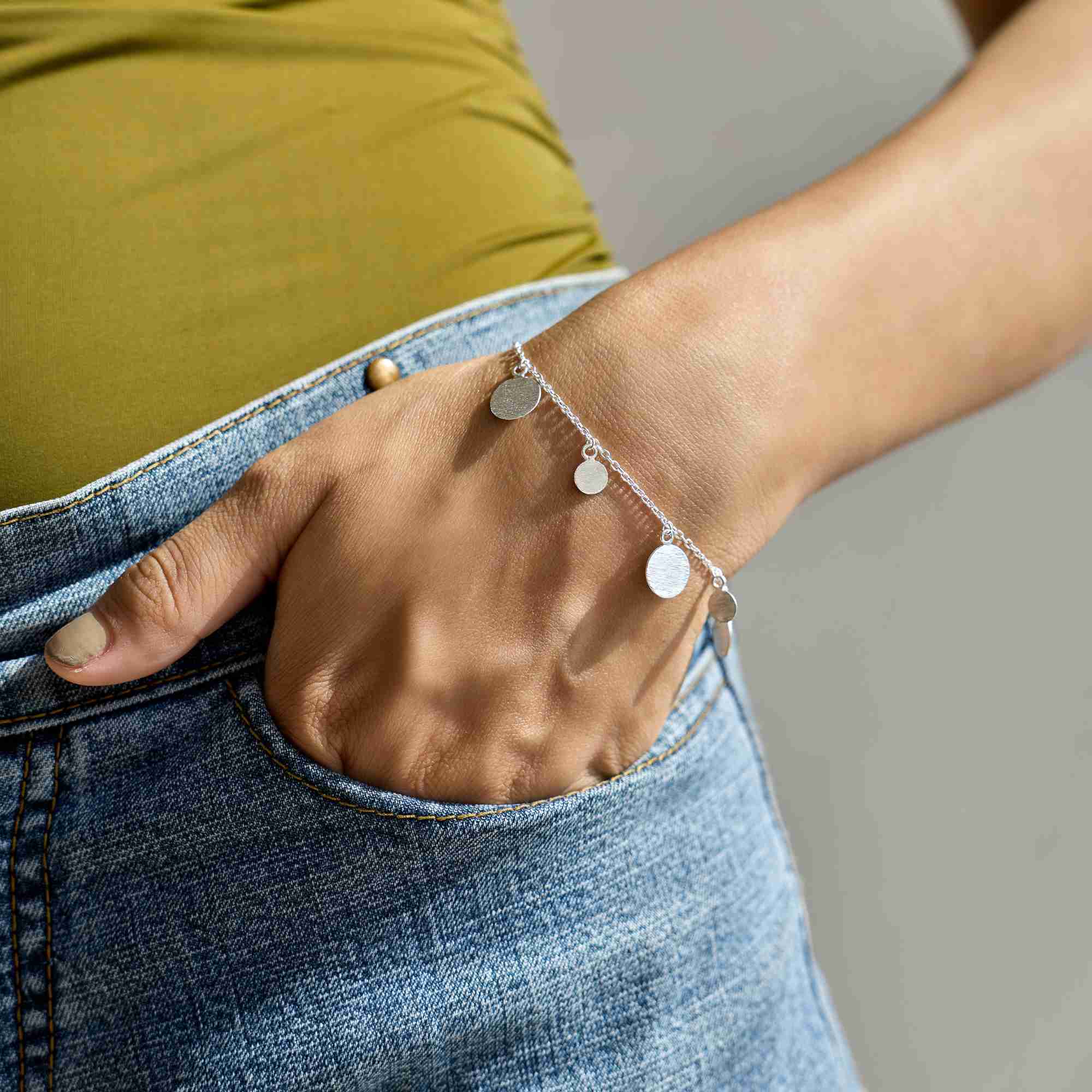 Model posing with hand in denim jeans pocket, wearing the silver Shimmering Disks Delicate Chain Bracelet with coin charm accents.