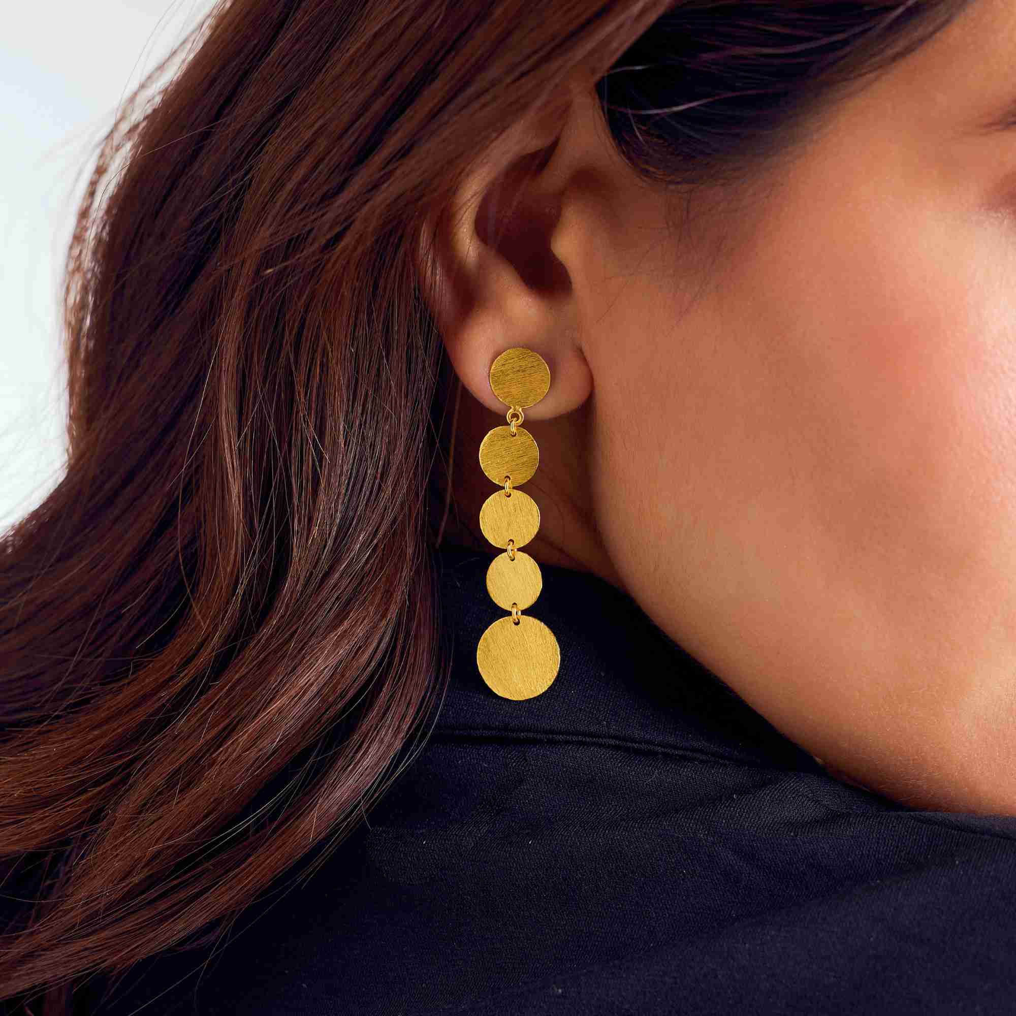 Close-up profile view showing a model wearing gold Shimmering Disks Graduating Disc Cascade Statement Earrings against a dark top, showcasing five linked brushed circular discs.