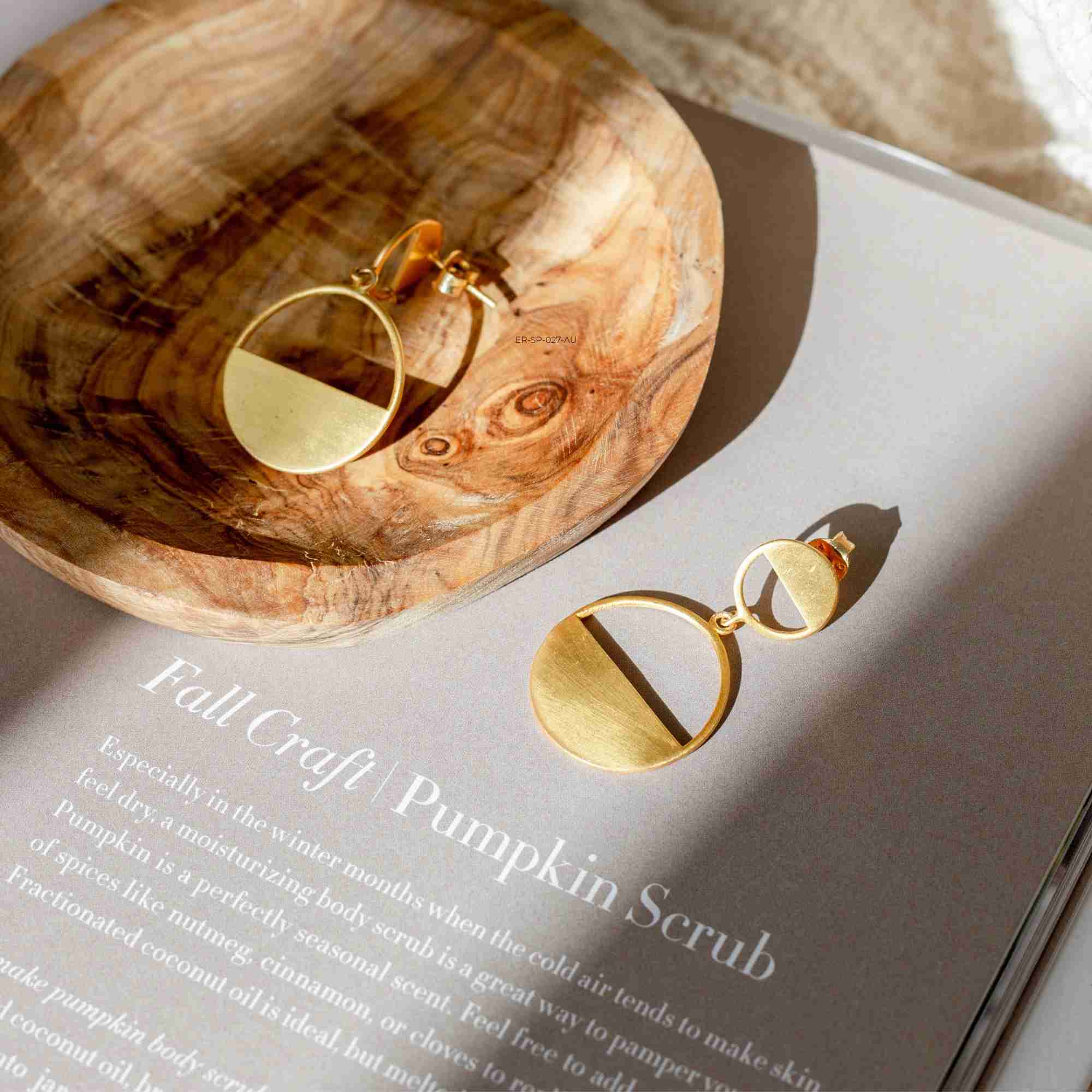 Studio product shot of Shimmering Disks Horizon Duo Drop Statement Earrings Gold arranged flat on a light surface, highlighting the linear drop design of a smaller top circle connected to a larger horizontal bar crescent disc with a signature hand-brushed matte gold texture.