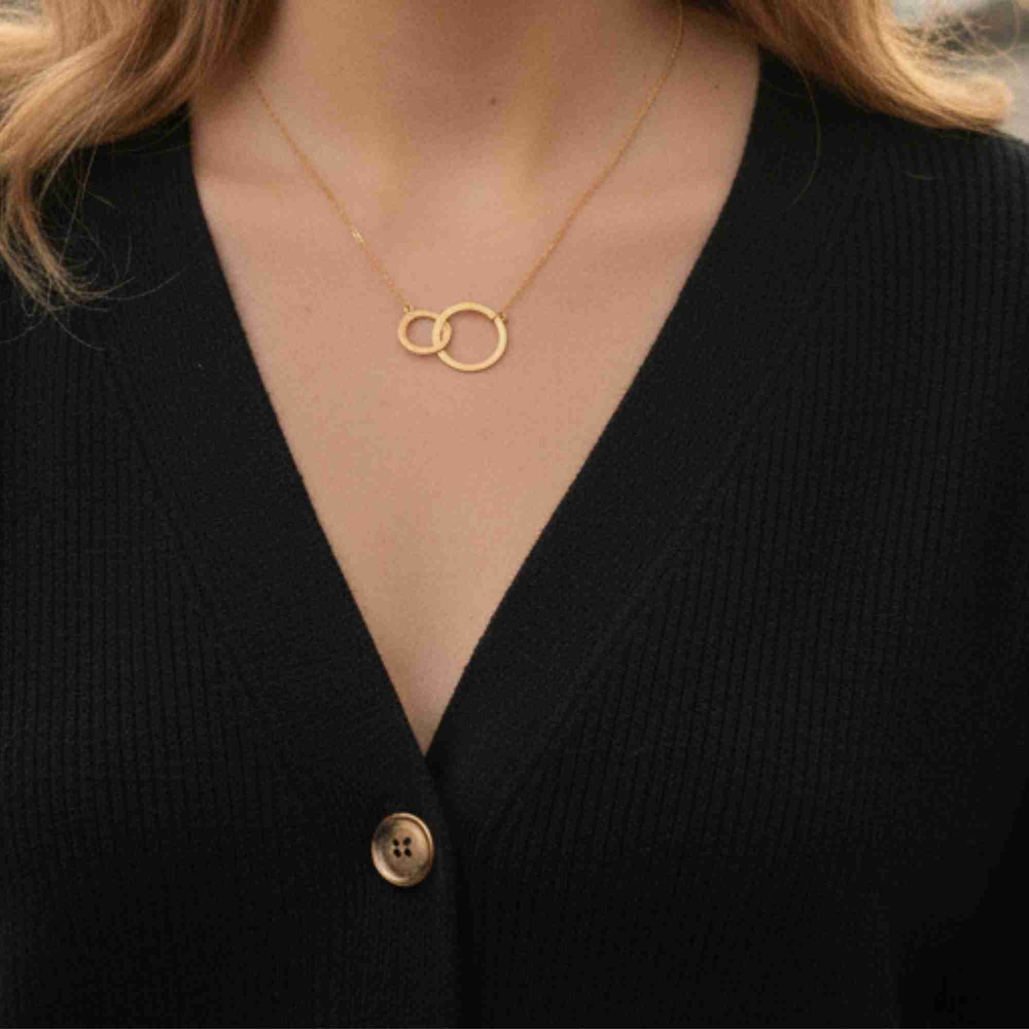 Close-up model view named Shimmering Disks (61).jpg showing a woman wearing the Shimmering Disks Interlinked Circles Necklace Gold over a black V-neck ribbed cardigan, highlighting the two delicate, interlocking gold geometric loops on a fine chain by Protsaah Zurich.