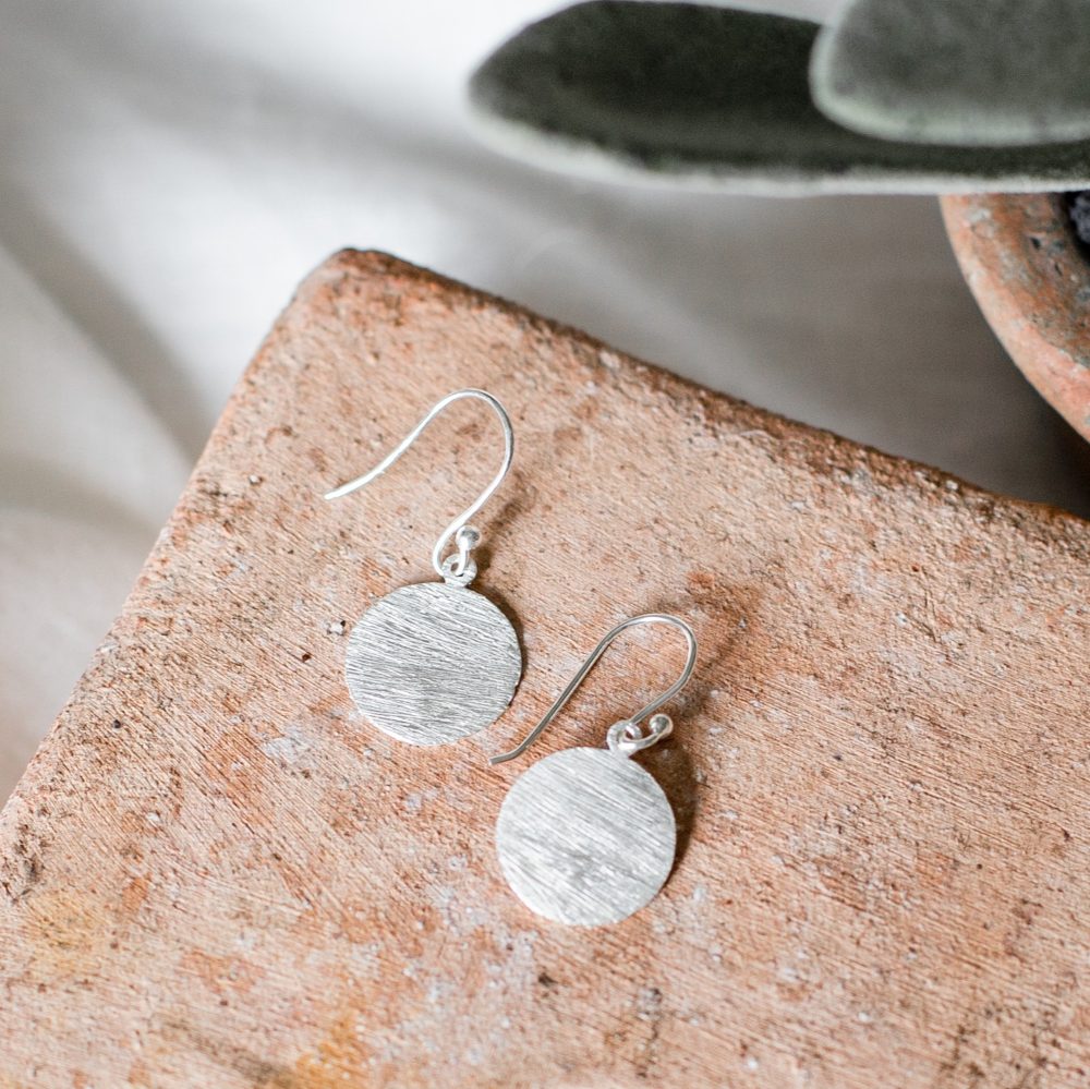 Disc drop earrings in silver with a hand-brushed finish lying on a terracotta tile