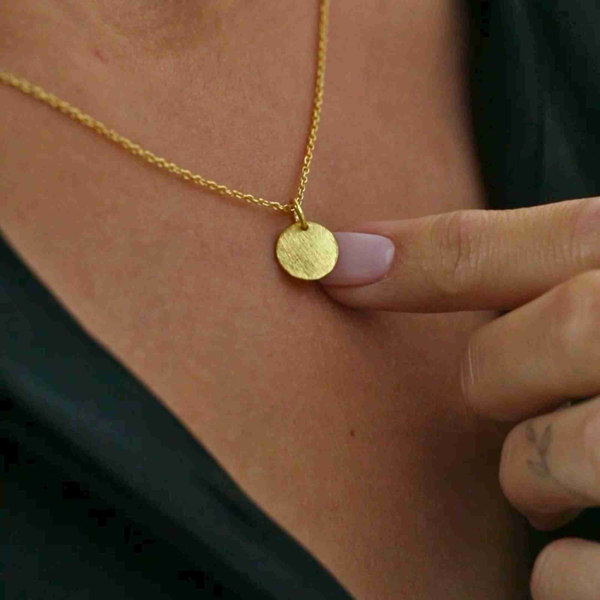 Close-up shot of a hand gently holding the round gold coin pendant on the Shimmering Disks Minimalist Pendant Necklace against black clothing, highlighting its textured surface.