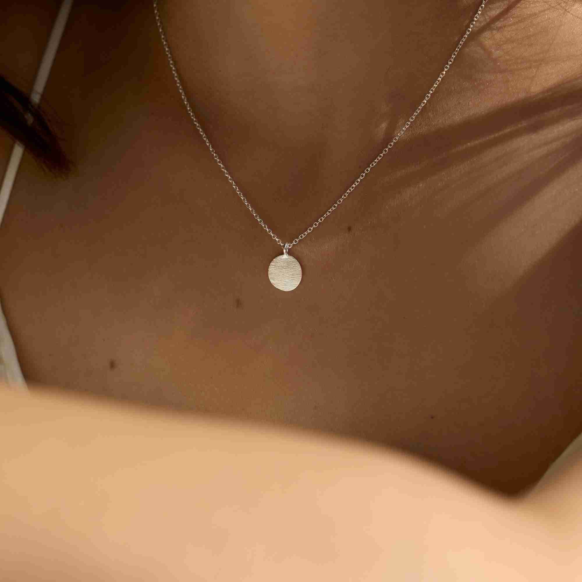 Close-up detail shot highlighting the textured, hand-brushed finish of the circular silver coin pendant on its delicate link chain.