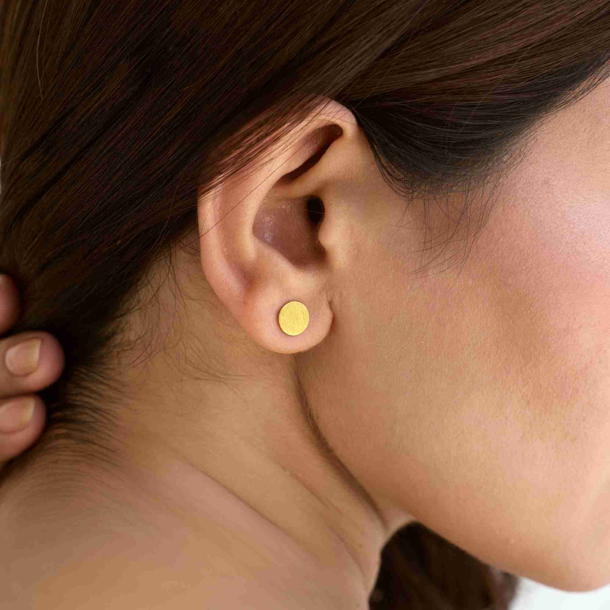 Side profile close-up of a model wearing the Shimmering Disks Minimalist Stud Earrings, displaying the hand-applied brushed-satin scratch texture on the gold circular disc.