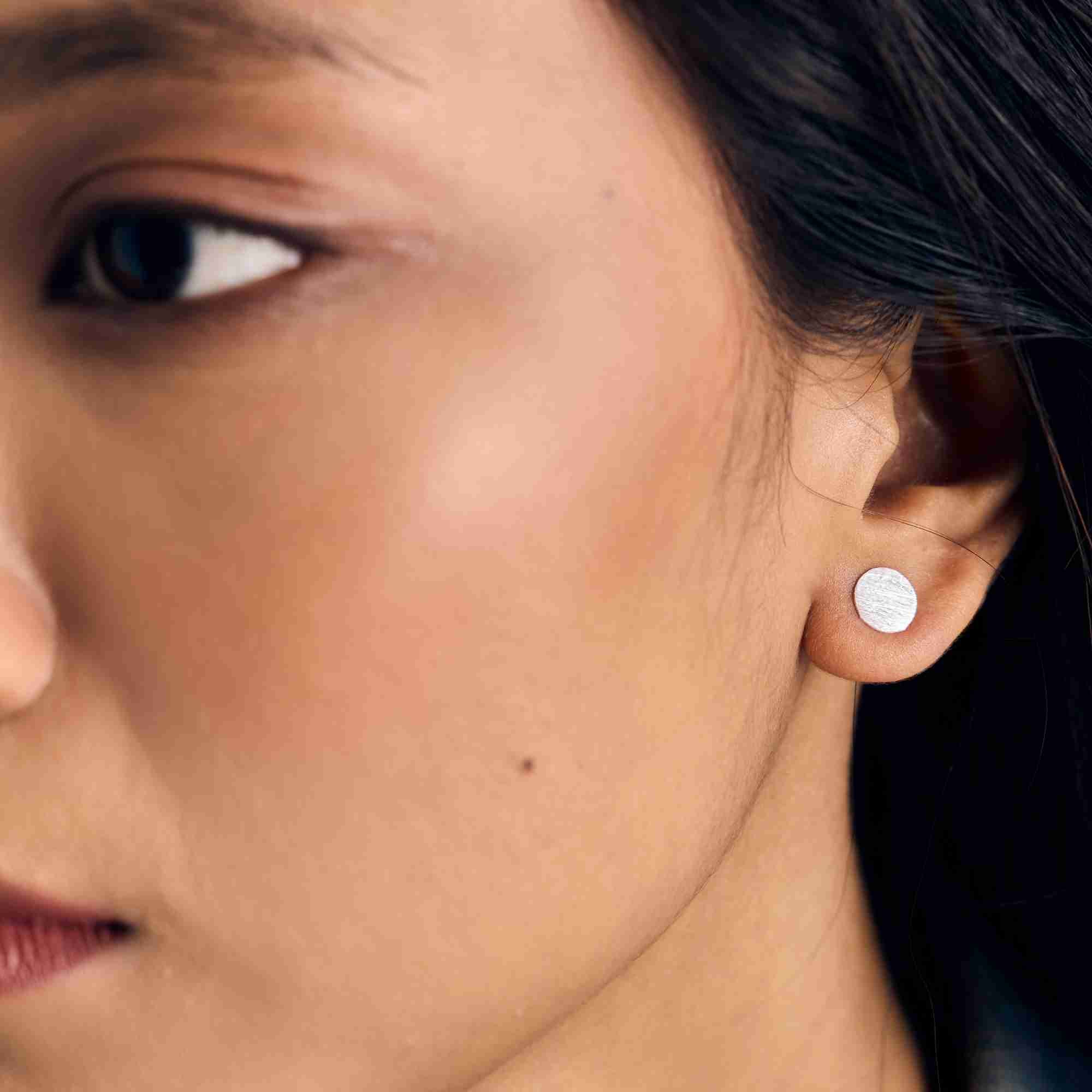Close-up portrait of a model wearing the Shimmering Disks Minimalist Stud Earrings in sterling silver, highlighting the 8.2 mm flat disc design on the earlobe.