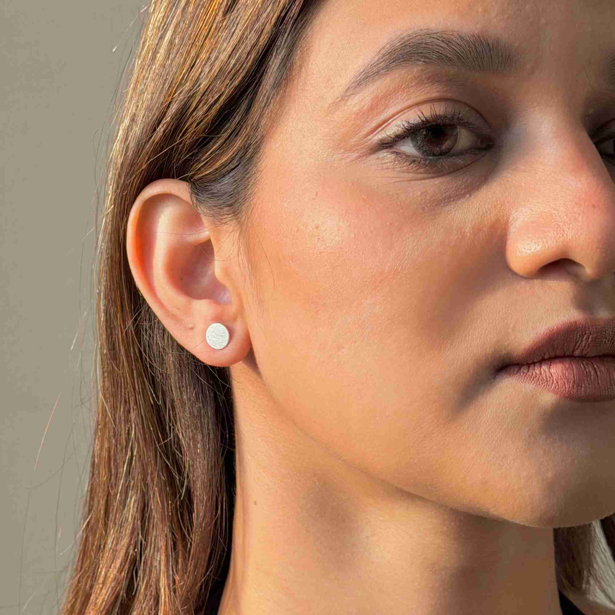 Model wearing the silver Shimmering Disks Minimalist Stud Earrings, displaying the brushed-satin scratch finish on a small circular stud.