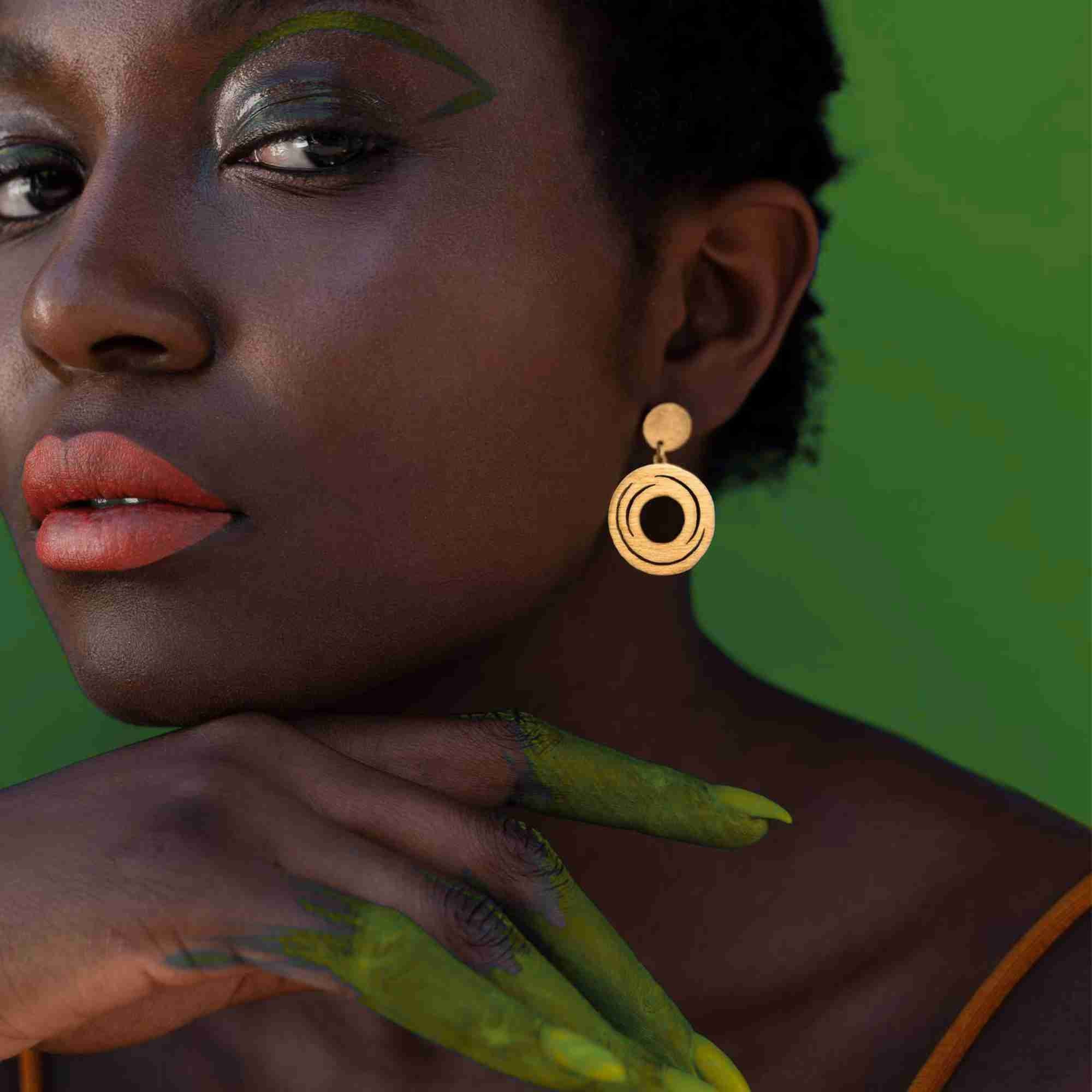 Model against a green backdrop wearing the gold Shimmering Disks Orbital Swirl Drop Statement Earrings, showcasing the layered concentric swirl pendant design.