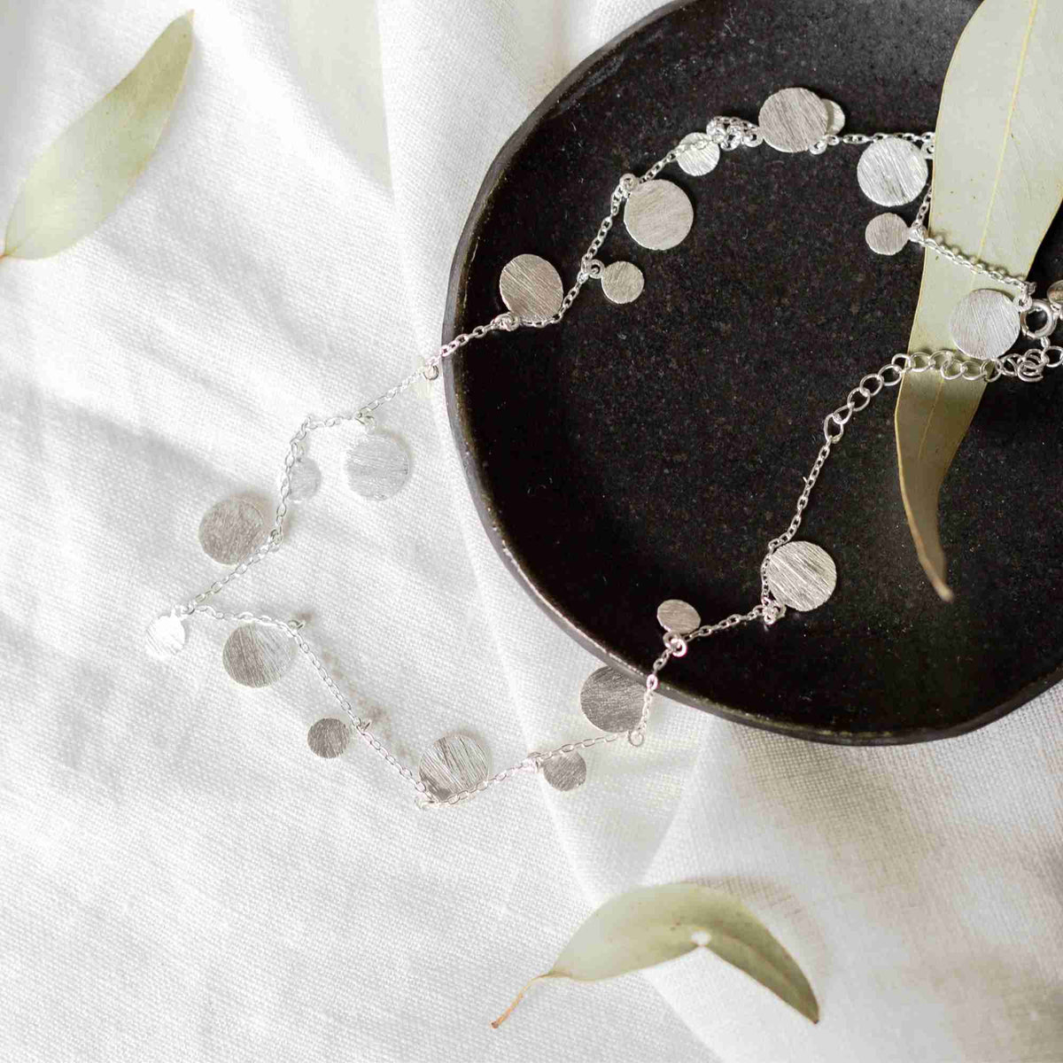 Silver scattered disc necklace with hand-brushed discs laid across white linen and a dark ceramic plate
