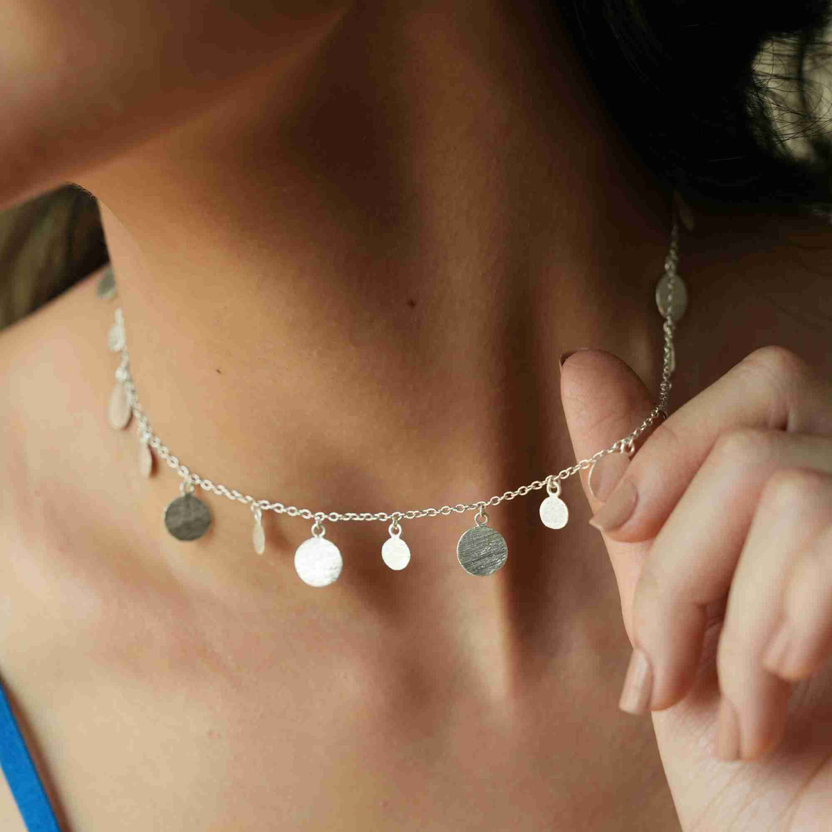Model wearing scattered disc necklace in silver at the base of the neck, lifting the chain with her fingers
