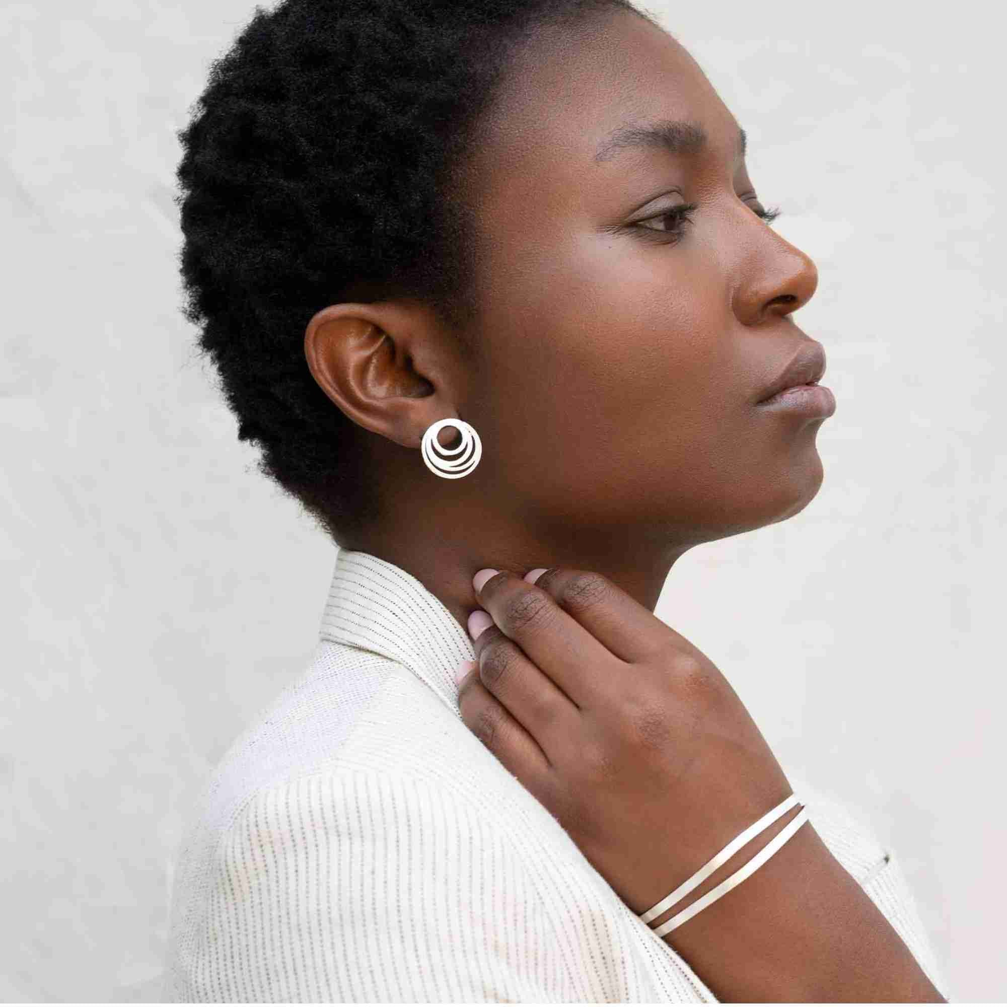 Profile shot of a model wearing a white pinstripe blazer, styled with the silver Shimmering Disks Sculptural Flat-Band Double Cuff on her wrist and matching concentric circle stud earrings.