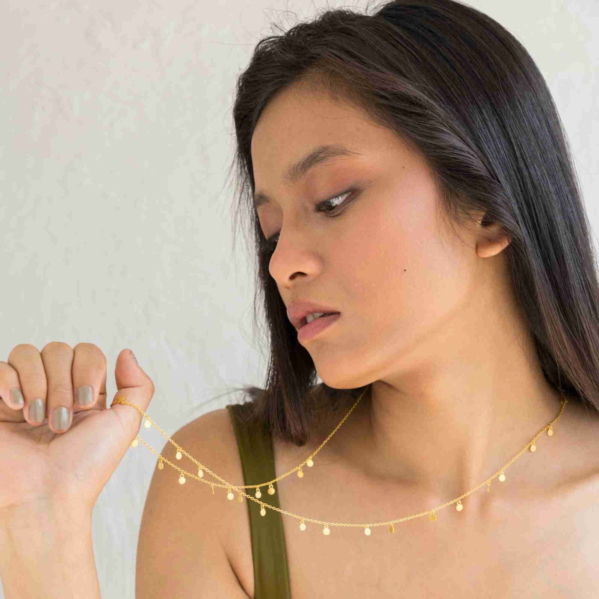 A lifestyle close-up of a woman model holding the Shimmering Disks Constellation Disc Chain Necklace Gold in her mouth, displaying the delicate gold link chain and hanging mini disc details against a textured white background.