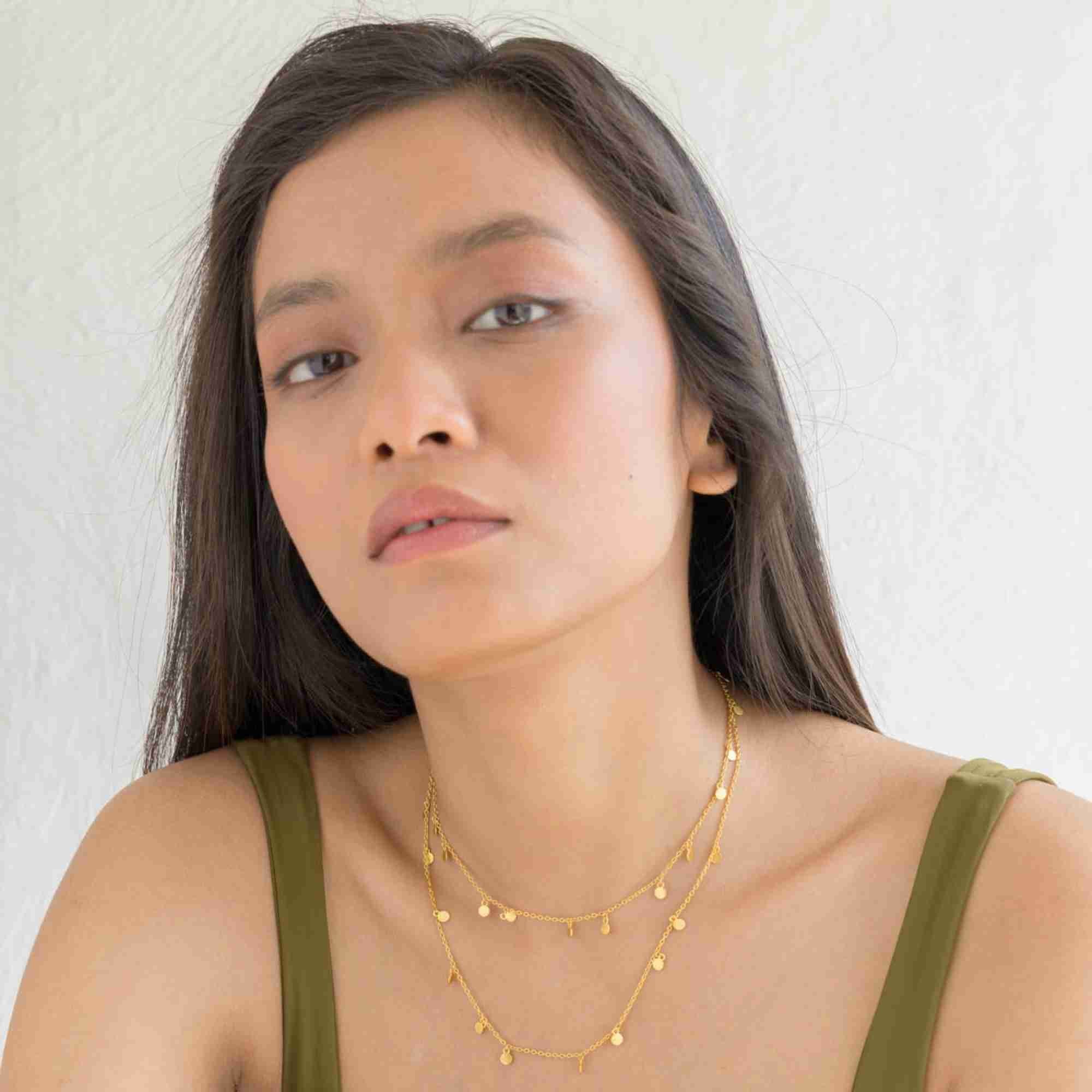 A lifestyle close-up of a woman model holding the Shimmering Disks Constellation Disc Chain Necklace Gold in her mouth, displaying the delicate gold link chain and hanging mini disc details against a textured white background.