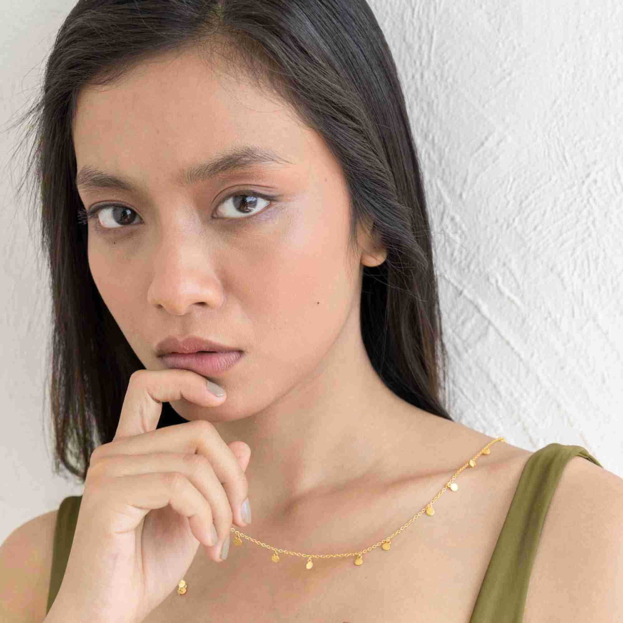 A lifestyle close-up of a woman model holding the Shimmering Disks Constellation Disc Chain Necklace Gold in her mouth, displaying the delicate gold link chain and hanging mini disc details against a textured white background.